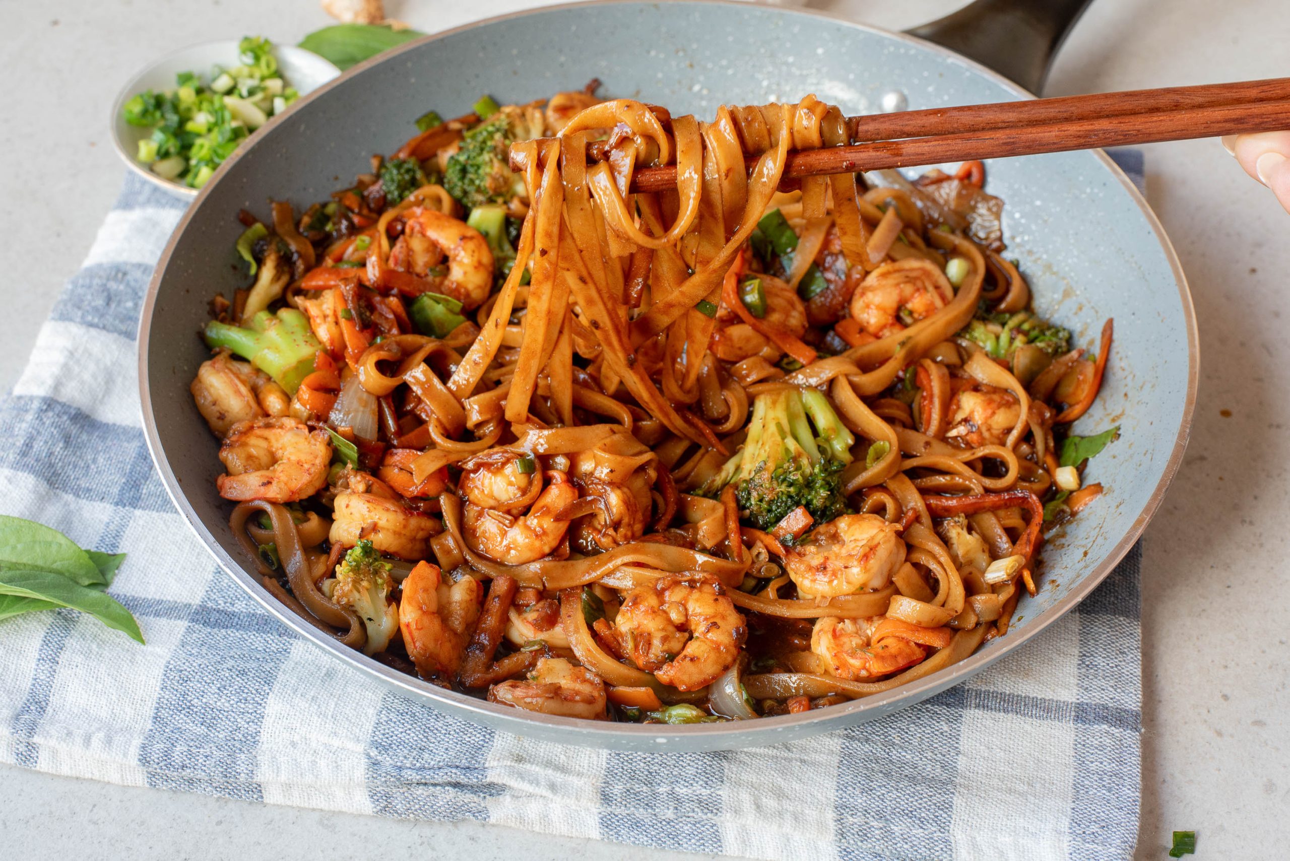 A pan of drunken Thai noodles with shrimp, broccoli, and vegetables is being served with chopsticks on a blue and white cloth.