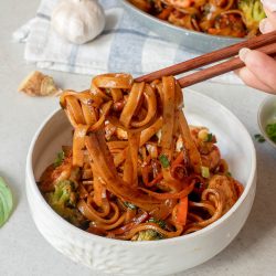A hand holding chopsticks lifts noodles from a white bowl filled with stir-fried noodles, vegetables, and sauce. A skillet with more noodles and a head of garlic are in the background.