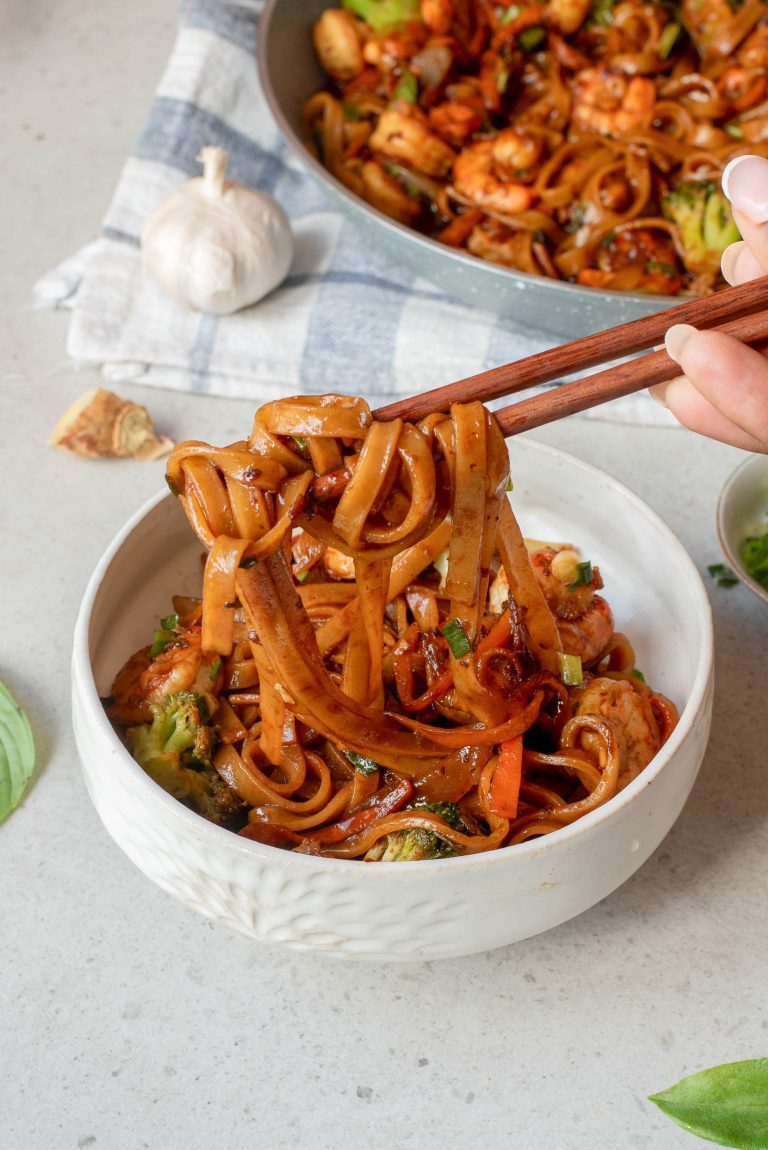 A hand holding chopsticks lifts noodles from a white bowl filled with stir-fried noodles, vegetables, and sauce. A skillet with more noodles and a head of garlic are in the background.
