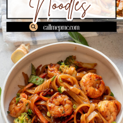 A bowl of Drunken Thai Noodles with shrimp, broccoli, and green onions sits on a table, accompanied by chopsticks and a pan of extra noodles in the background.