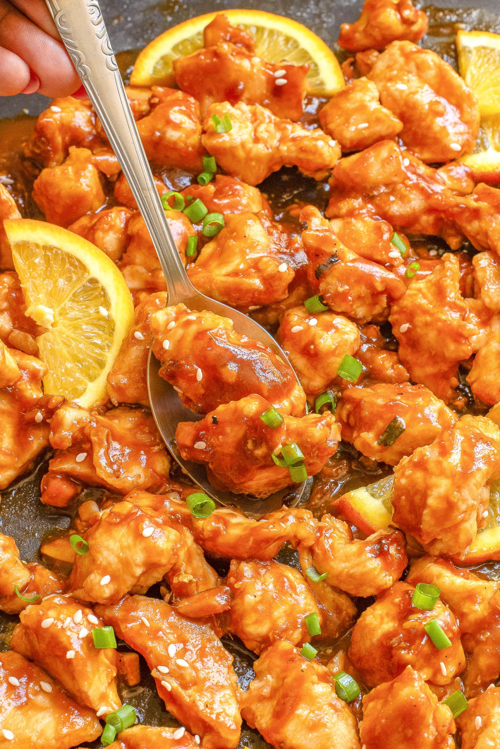 Close-up of orange chicken garnished with sesame seeds, chopped green onions, and orange slices, with a spoon serving a portion.