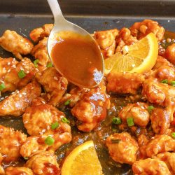 A spoon drizzles orange sauce over pieces of cooked chicken garnished with sesame seeds, chopped green onions, and orange slices on a baking sheet.