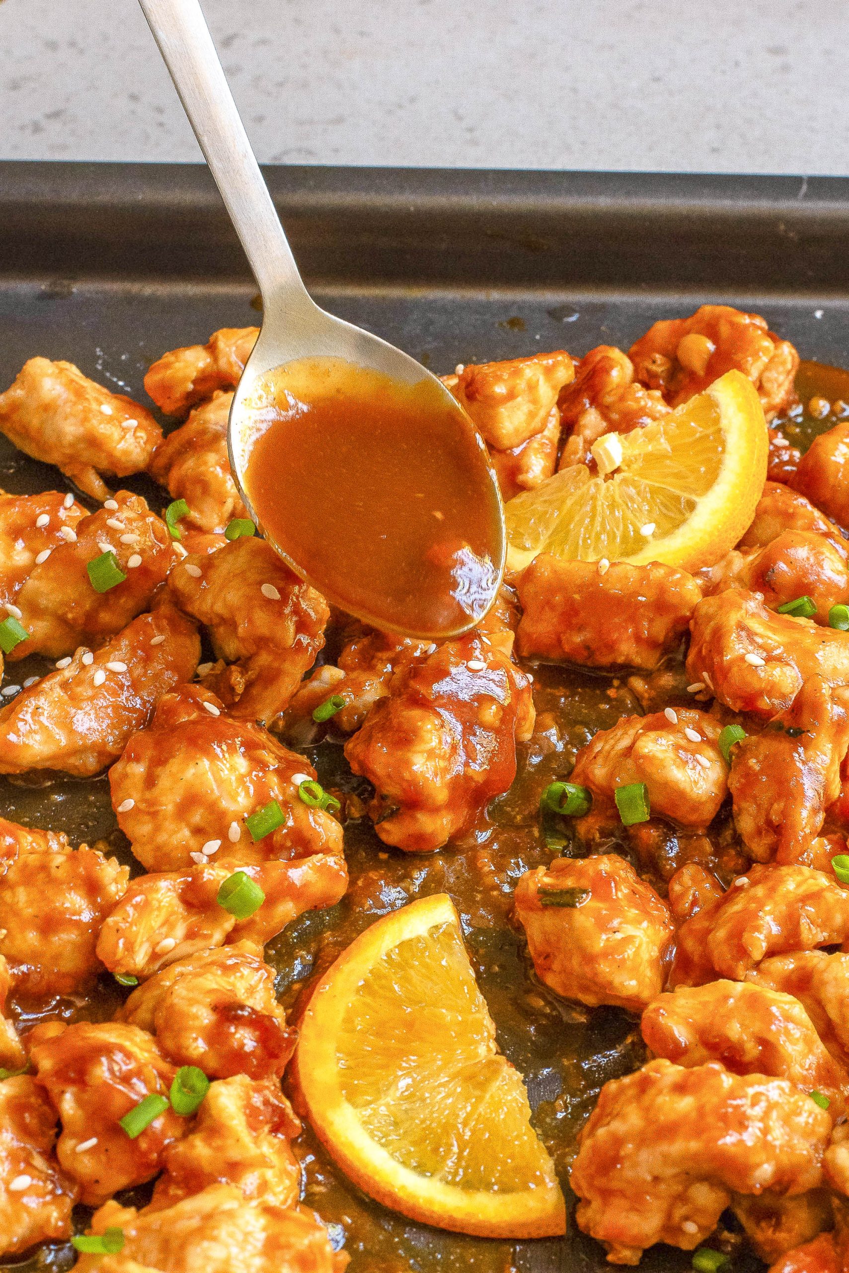 A spoon drizzles sauce over pieces of cooked protein garnished with sesame seeds and chopped green onions on a baking sheet.