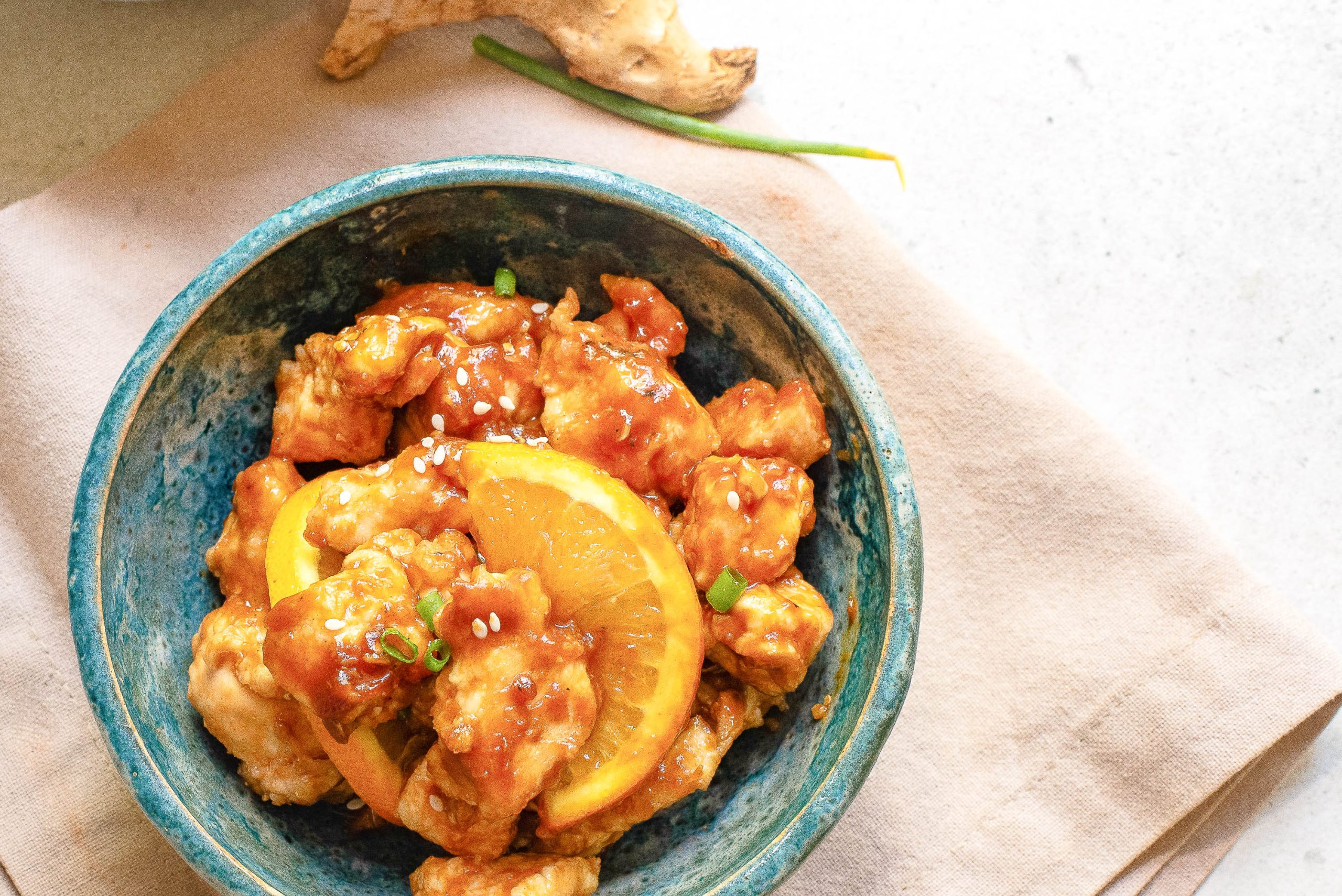 A blue bowl filled with orange chicken topped with sesame seeds and garnished with orange slices, placed on a beige napkin.