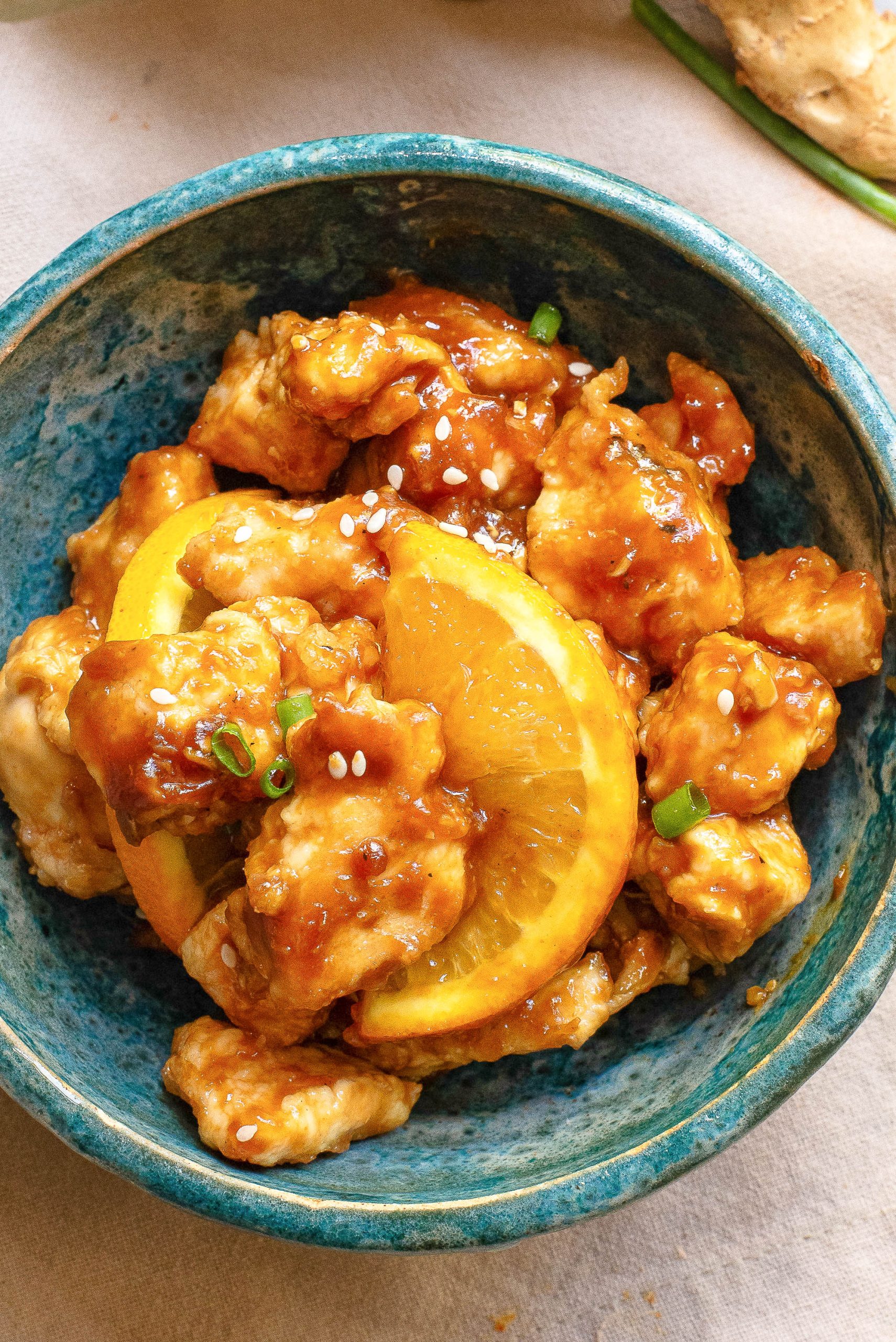 A blue bowl filled with orange chicken garnished with sesame seeds, green onions, and two orange slices.