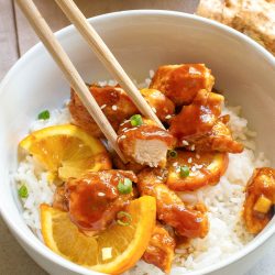 A bowl of white rice topped with orange chicken pieces, orange slices, and chopped green onions, with chopsticks picking up a piece of chicken.