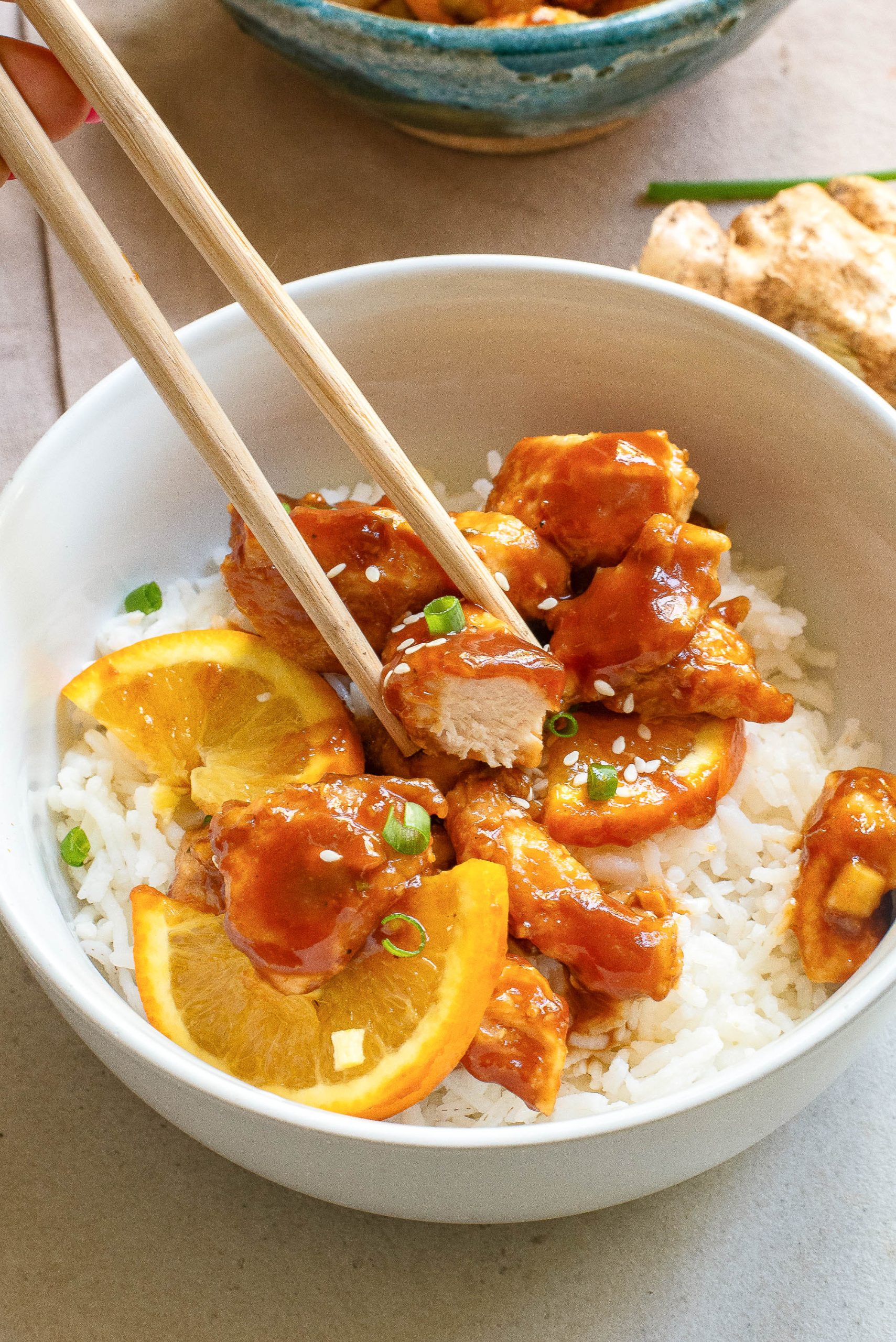 A bowl of white rice topped with orange chicken pieces, orange slices, and chopped green onions, with chopsticks picking up a piece of chicken.