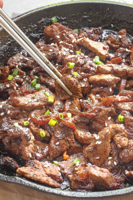 A close-up of cooked marinated beef slices in a pan, garnished with sesame seeds and chopped green onions, with metal chopsticks picking up a piece.