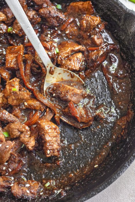 Chunks of cooked beef in a dark sauce with sliced vegetables in a cast iron skillet, stirred with a metal spoon.
