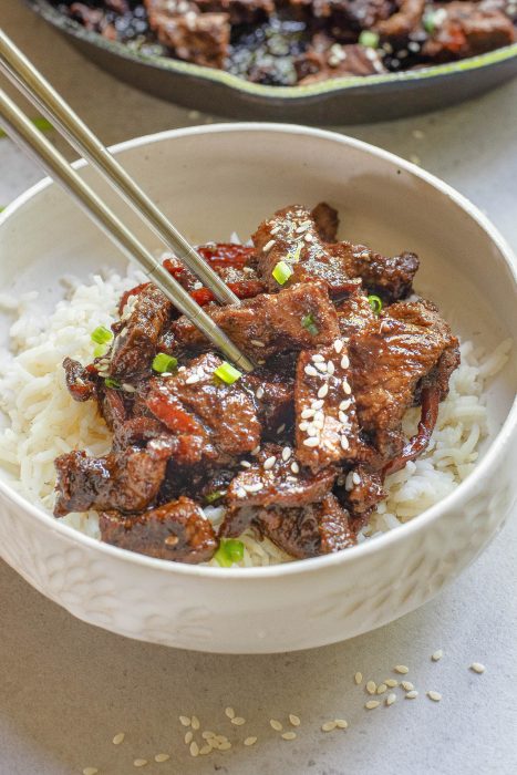 A bowl of white rice topped with cooked beef strips, garnished with sesame seeds and chopped green onions. Chopsticks are picking up a piece of beef.