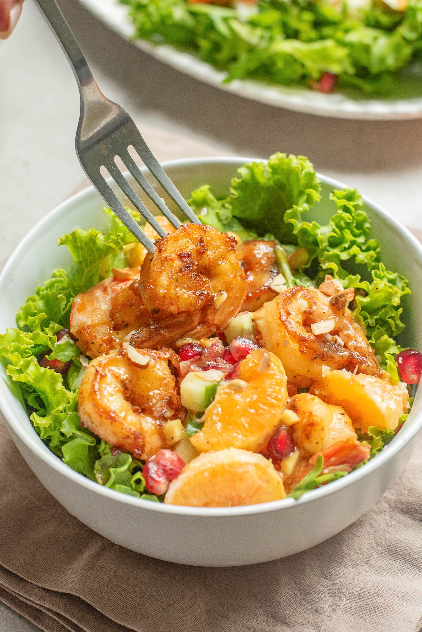 A fork holds a grilled shrimp over a salad with lettuce, orange slices, pomegranate seeds, and chopped vegetables in a white bowl.