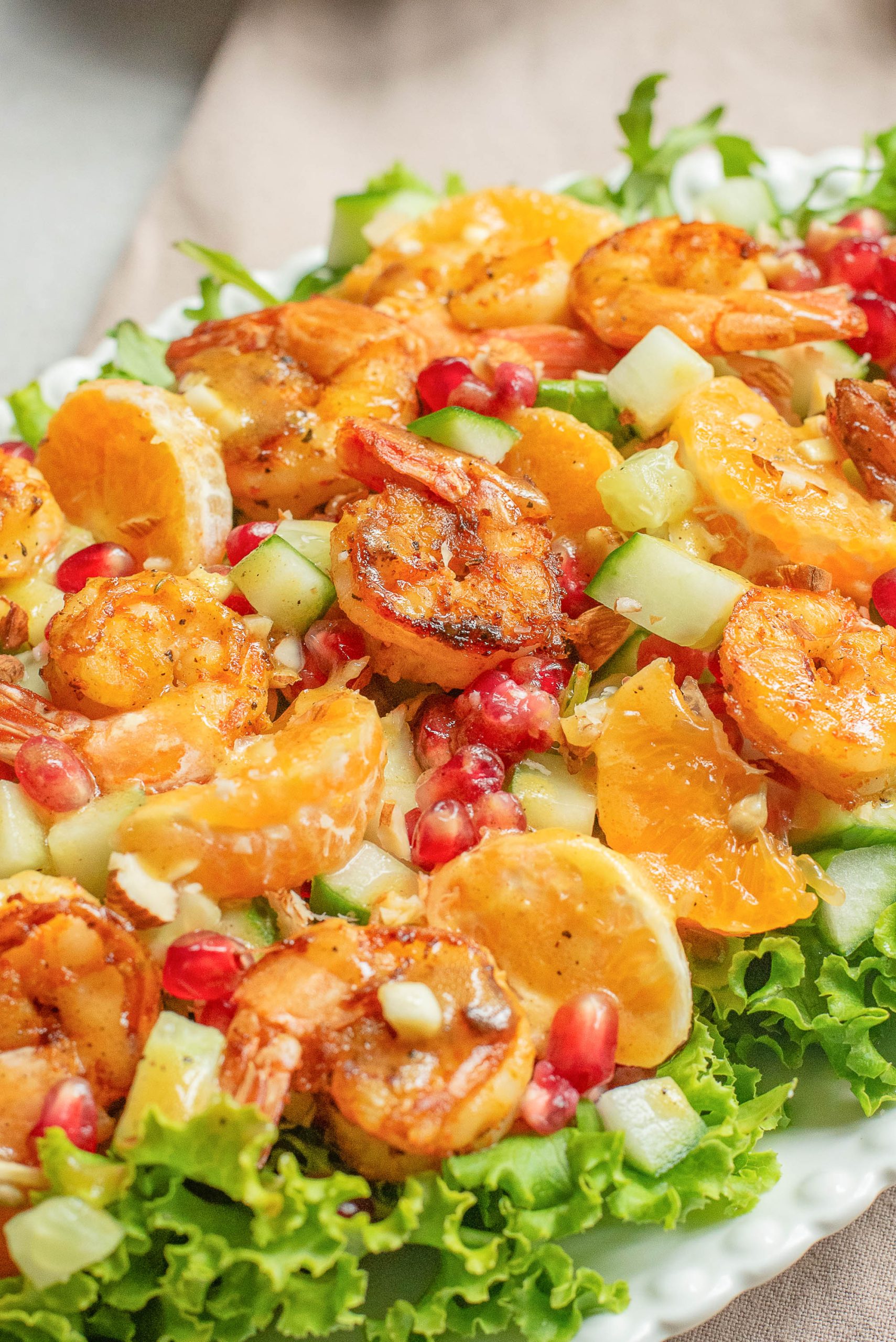 A plate of salad topped with grilled shrimp, orange slices, pomegranate seeds, cucumber pieces, and served on a bed of lettuce.
