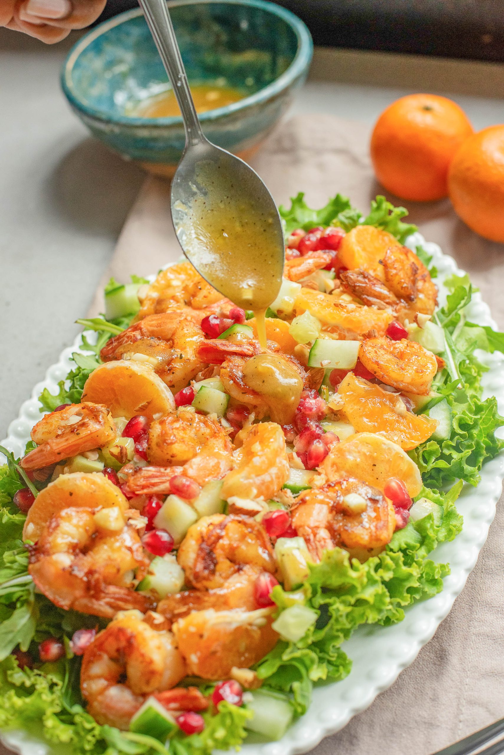 A platter of greens being drizzled with dressing from a spoon. A bowl and two whole oranges are in the background.