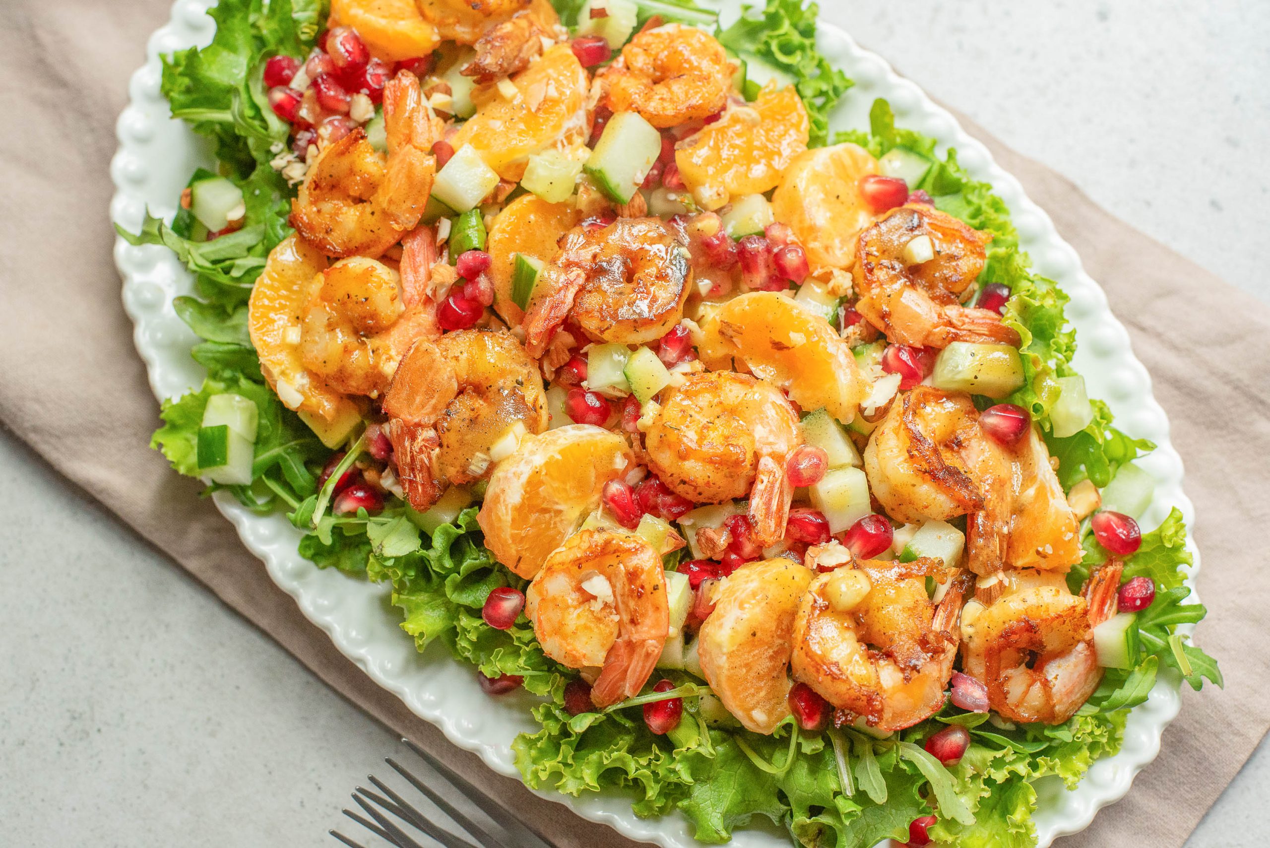 A plate of salad topped with cooked shrimp, orange slices, cucumber, pomegranate seeds, and lettuce on a white serving dish.