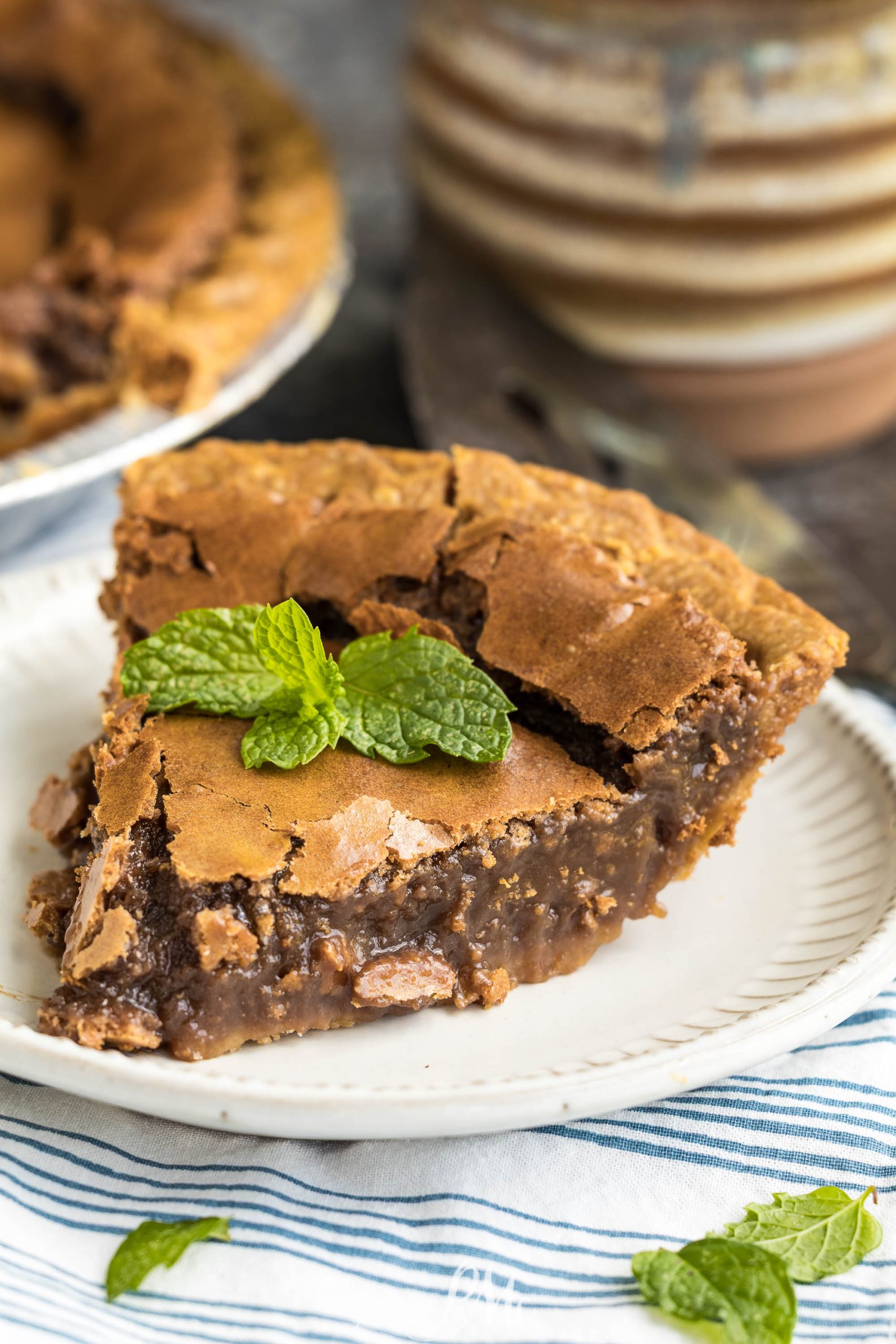 A slice of chocolate chess pie on a white plate, topped with fresh mint leaves, with a striped napkin underneath.