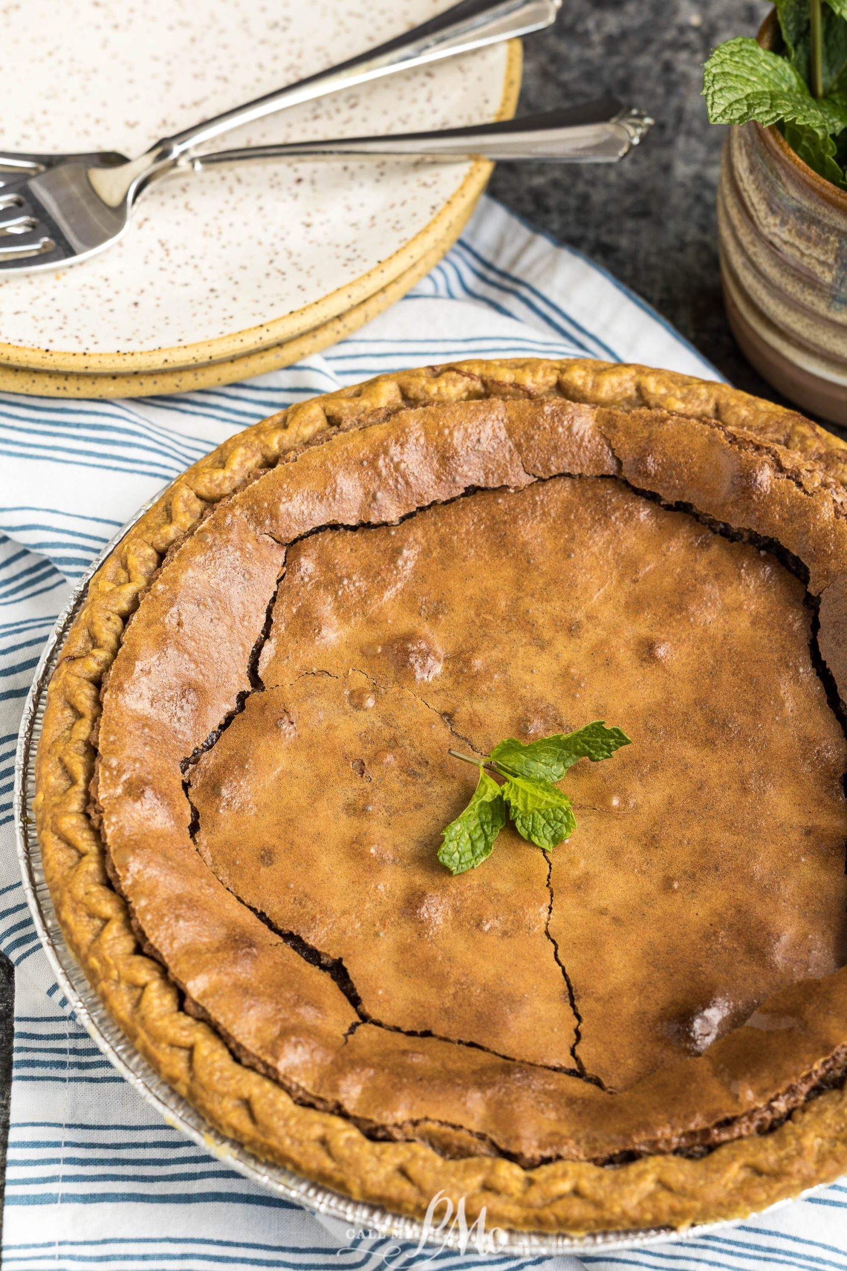 A baked chocolate pie with a cracked top, garnished with a sprig of mint, sits on a striped cloth next to two stacked plates and utensils.