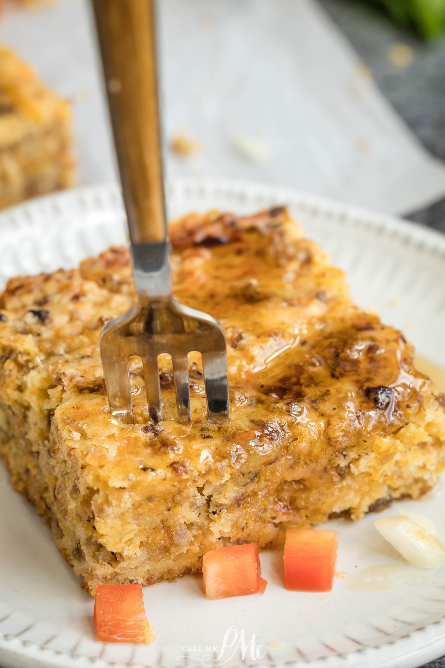 A fork pressed into a square piece of cornbread on a white plate, drizzled with honey and garnished with small diced tomato and onion.