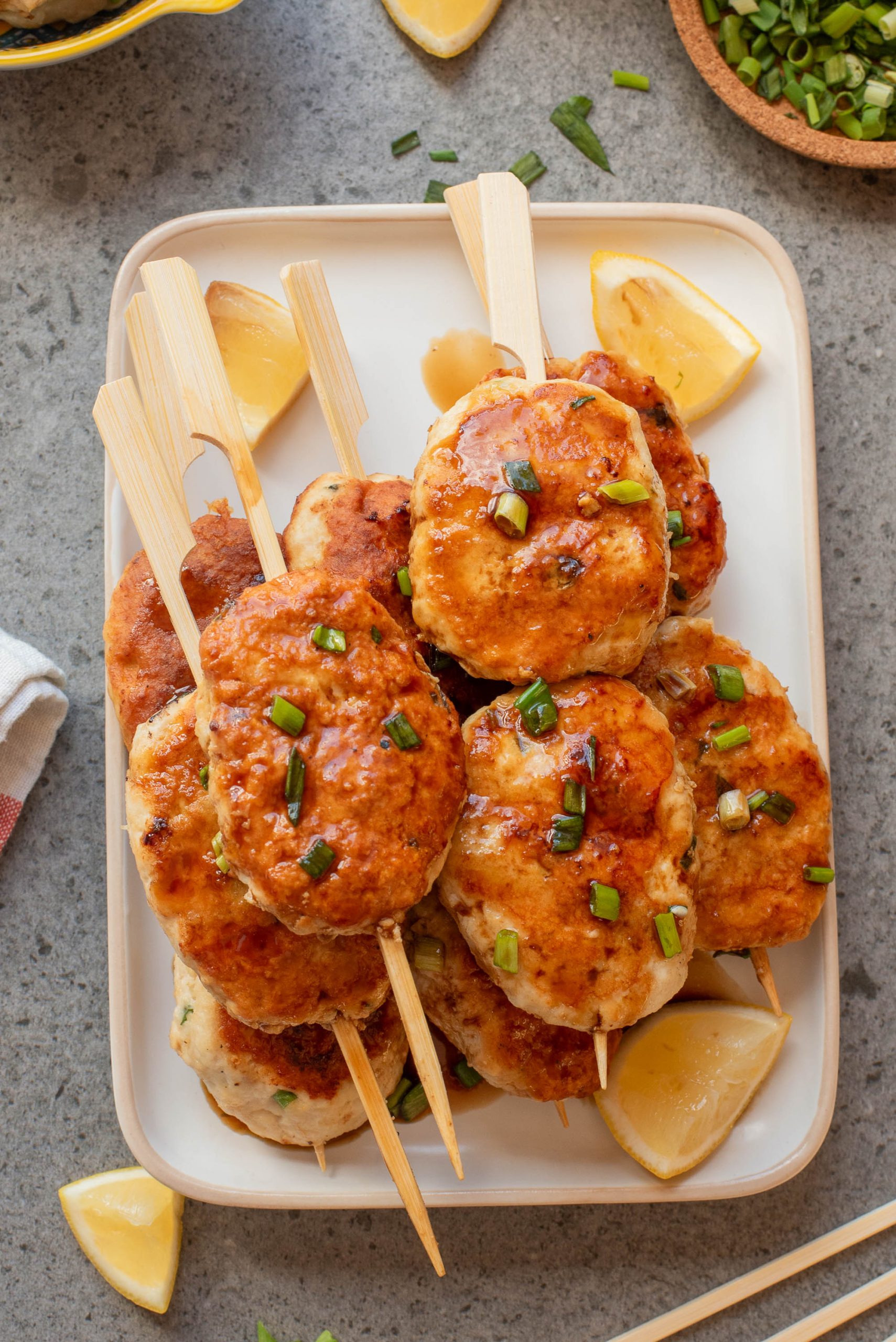 A plate of skewered grilled chicken patties garnished with chopped green onions, served with lemon wedges on a gray surface.