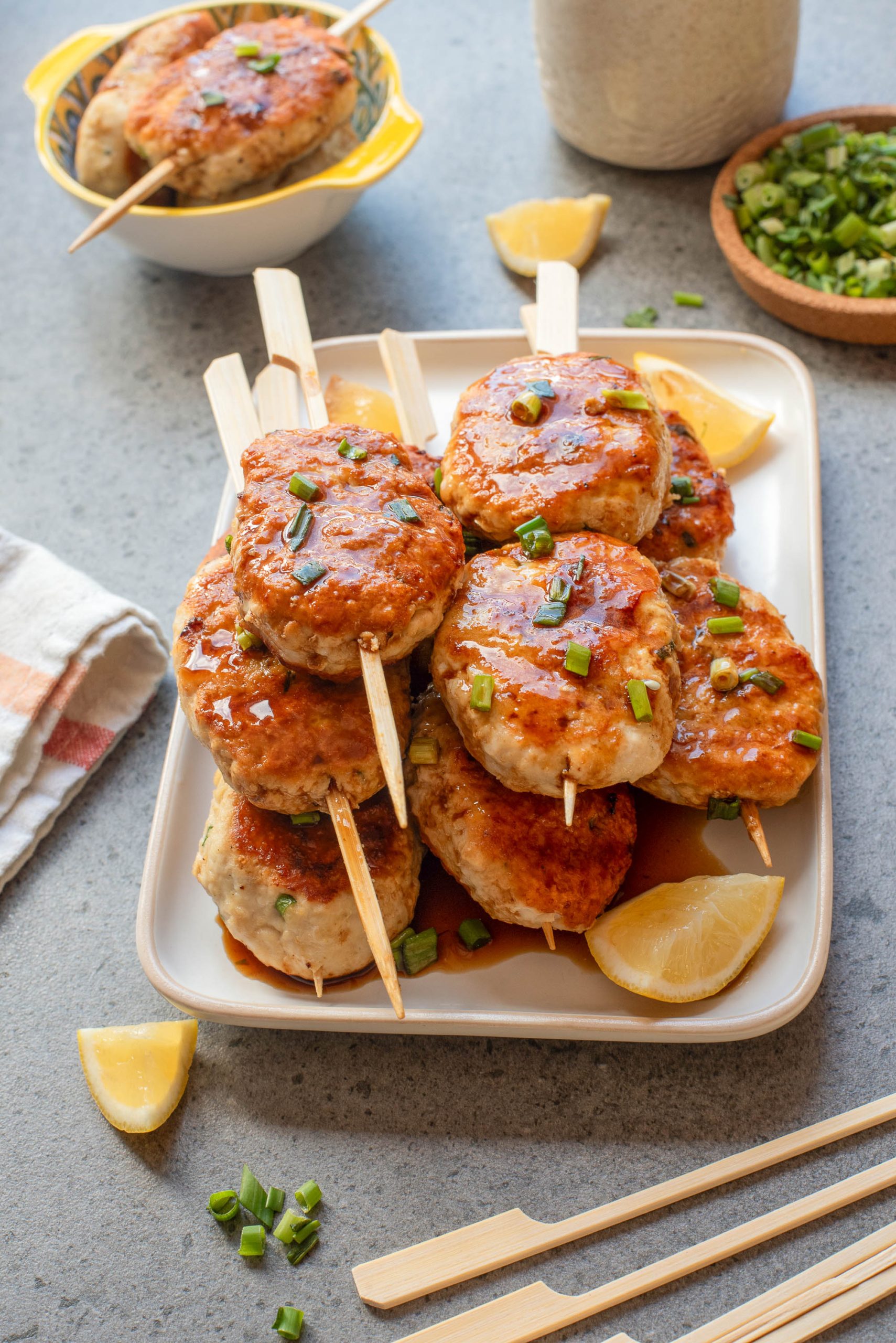 A plate of skewered Tsukune Japanese Meatballs topped with chopped green onions, served with lemon wedges on the side.