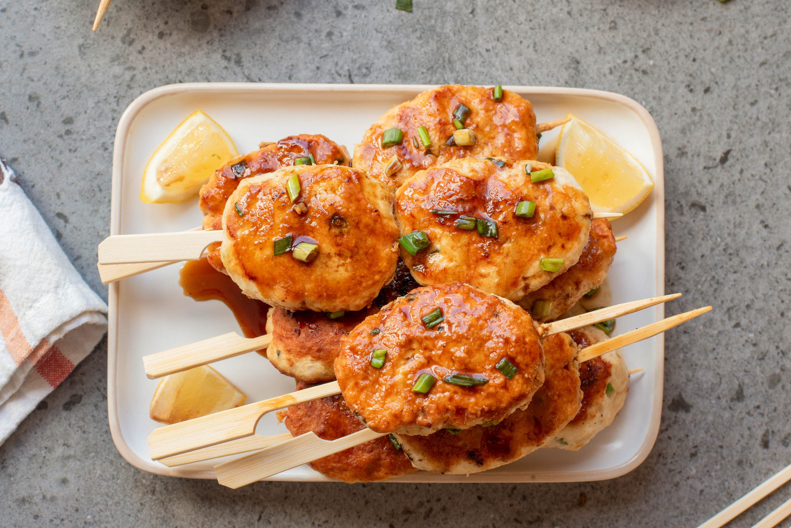 A plate of glazed chicken meatballs on wooden skewers, garnished with chopped green onions and served with lemon wedges.