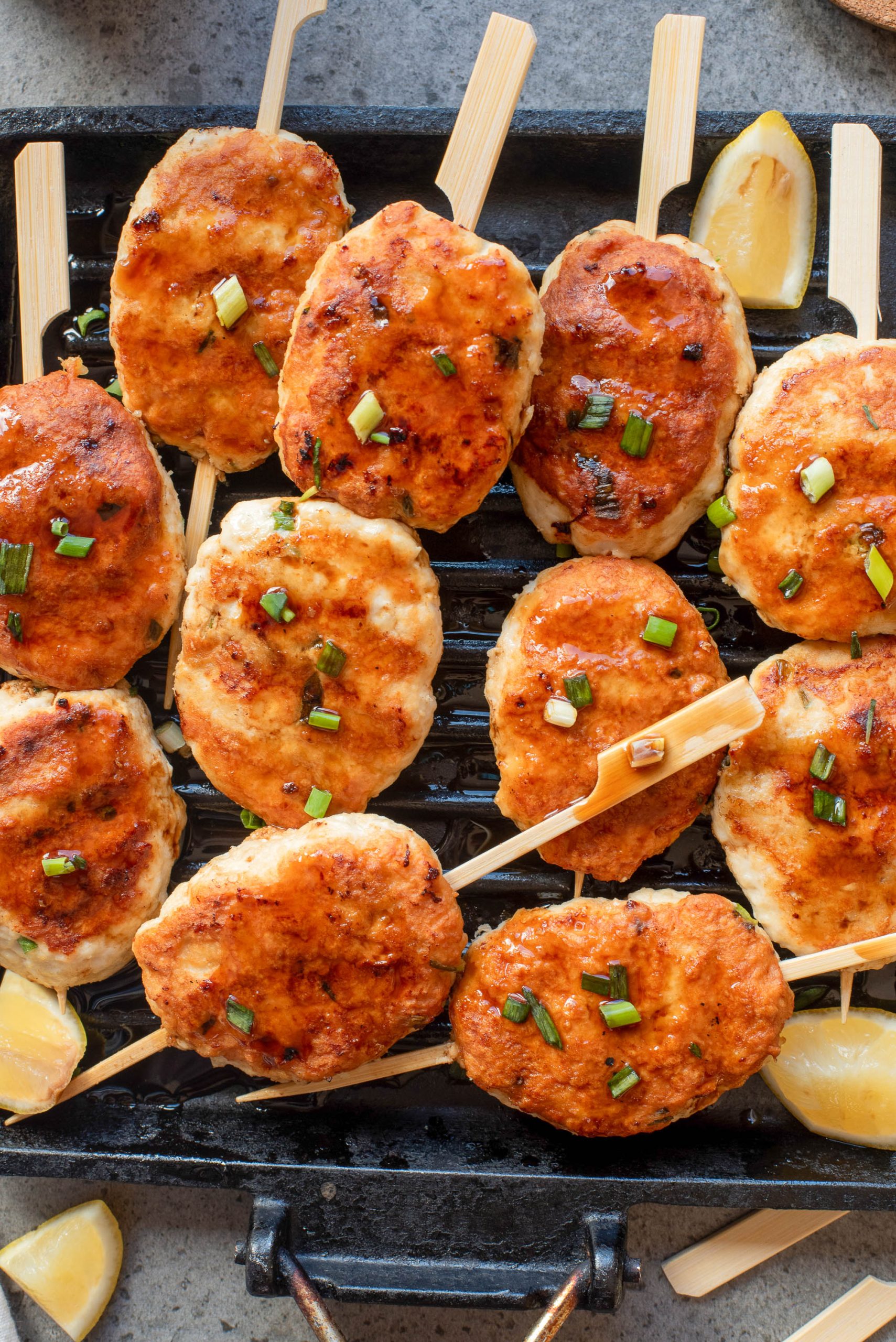 Grilled Tsukune Japanese Meatballs topped with chopped green onions are arranged on a grill pan, with lemon wedges placed on the sides.