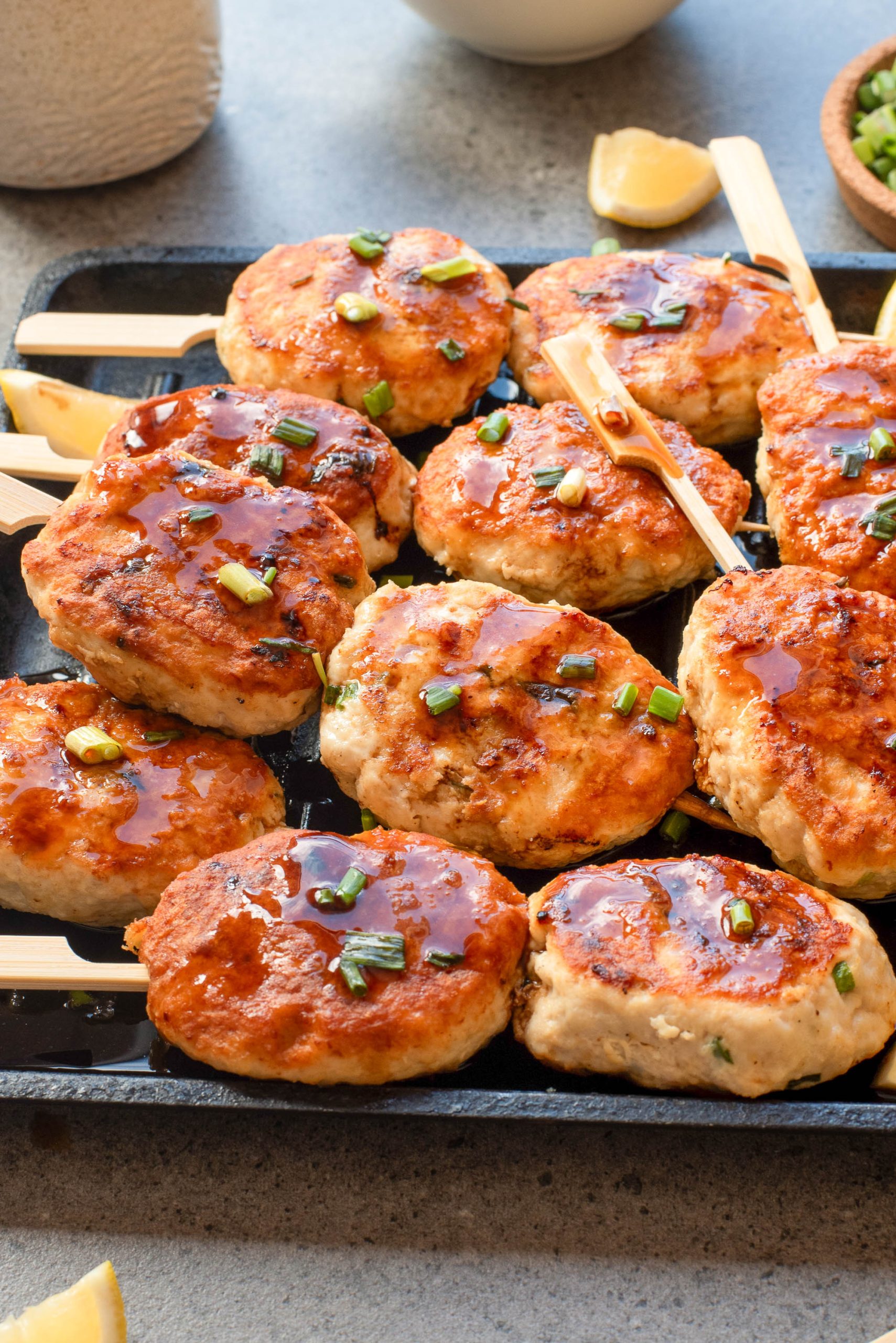 Grilled chicken patties on skewers topped with sauce and chopped green onions, served on a black tray with lemon wedges on the side.