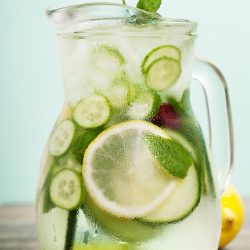 A glass pitcher filled with ice water, lemon slices, cucumber, mint leaves, and berries sits with condensation on the outside. Two striped straws and a lemon rest nearby—a refreshing idea for fruit-infused water recipes.