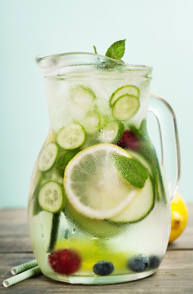 A glass pitcher filled with ice water, lemon slices, cucumber, mint leaves, and berries sits with condensation on the outside. Two striped straws and a lemon rest nearby—a refreshing idea for fruit-infused water recipes.