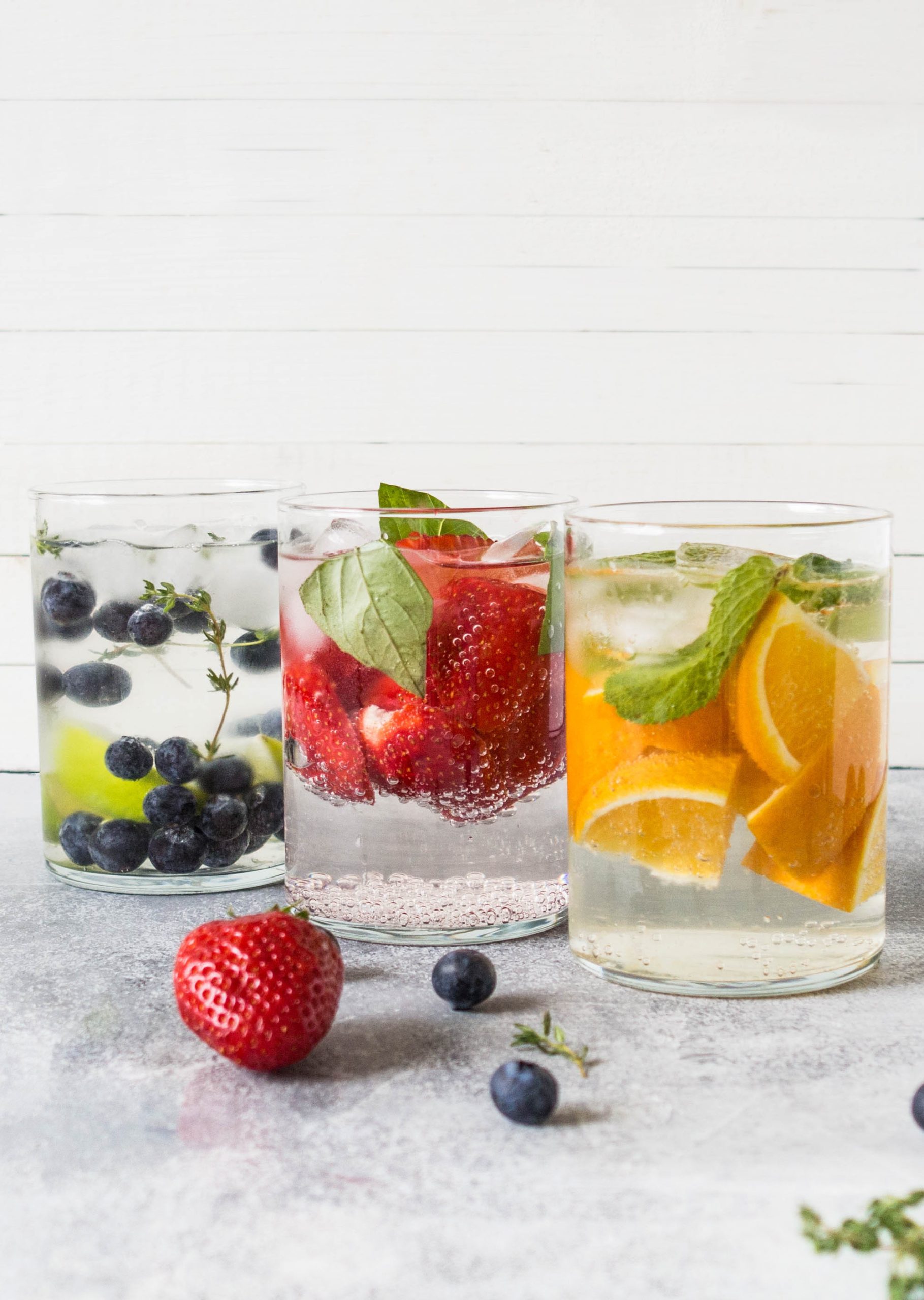 Three glasses of sparkling water infused with blueberries and thyme, strawberries and basil, and orange slices with mint showcase creative Fruit-Infused Water Recipes, with fresh fruits scattered on the table in front.