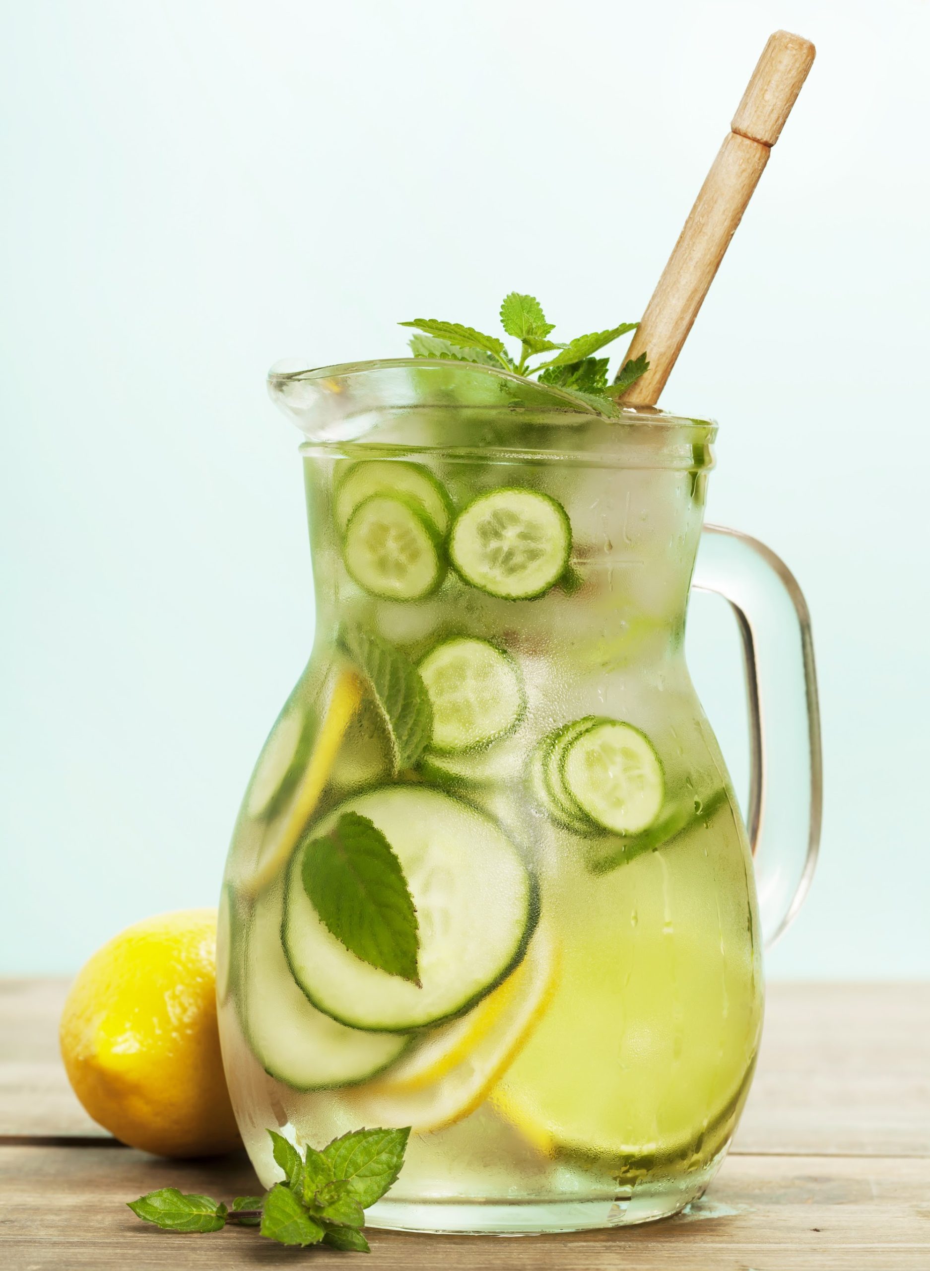 A glass pitcher of cucumber lemon water with mint leaves and a wooden stirrer sits on a wooden surface next to a whole lemon.