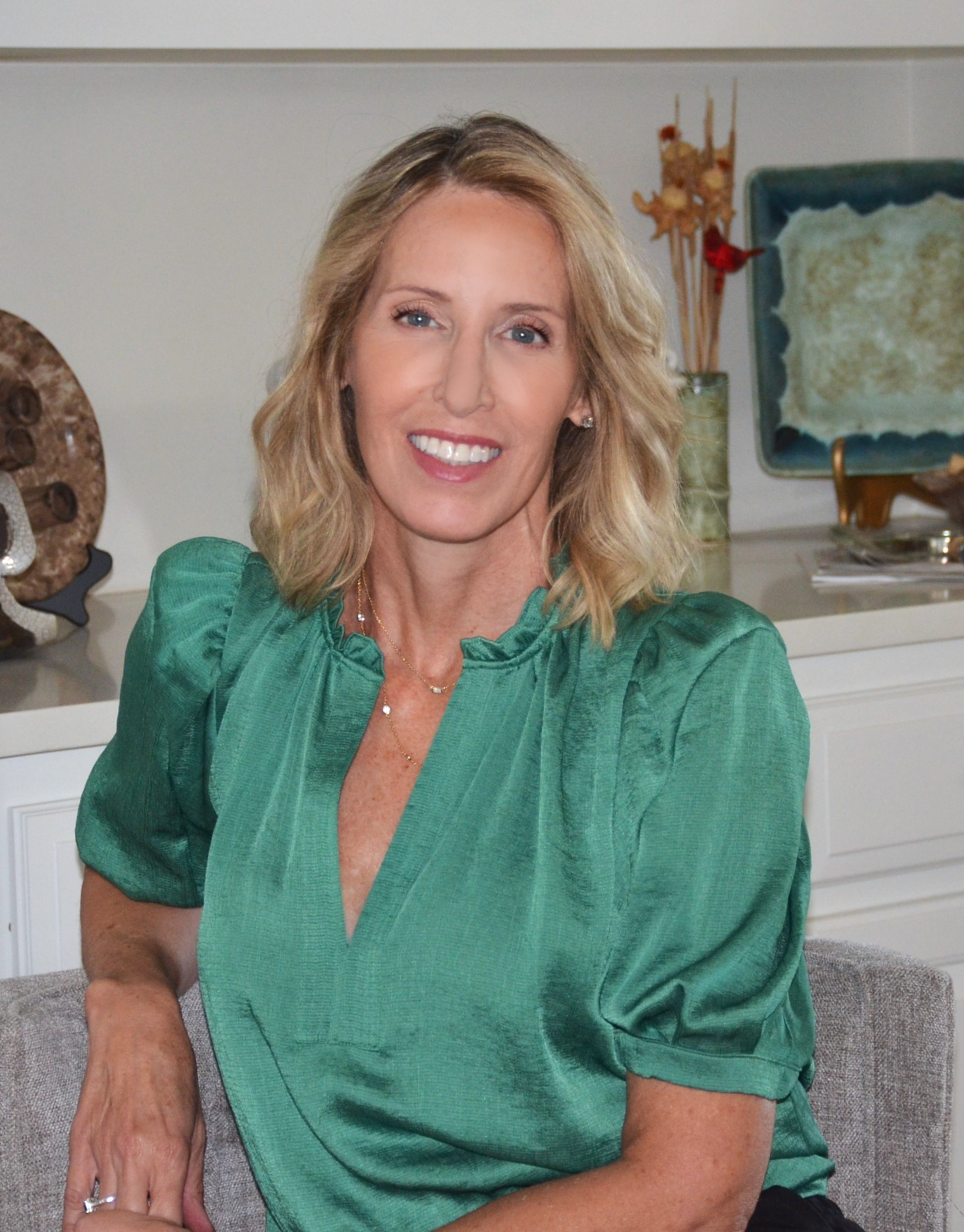 A woman with blonde hair wearing a green blouse sits on a gray chair and smiles at the camera; decorative items are on a shelf behind her.