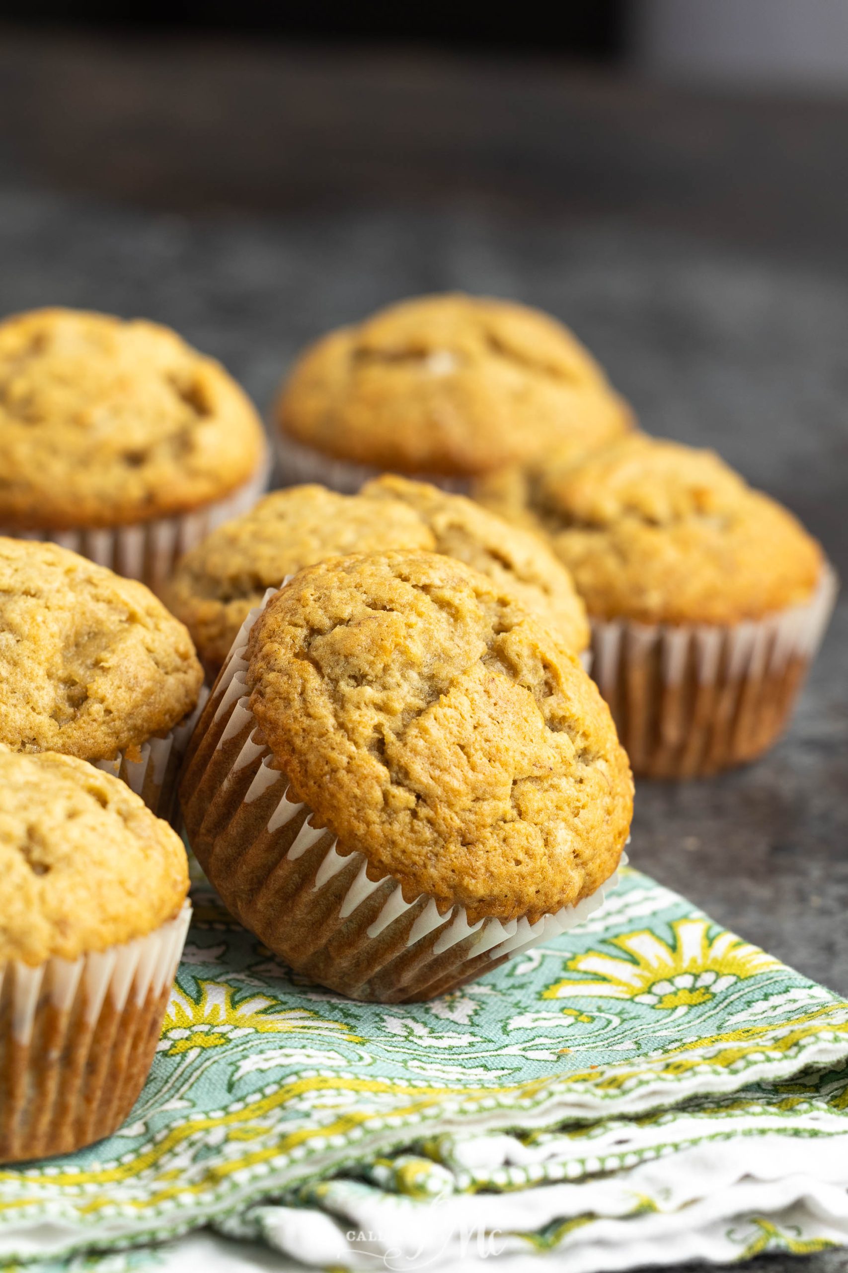A group of banana muffins in paper liners arranged on a patterned cloth with a dark background.