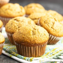 Several golden-brown muffins arranged on a patterned cloth, with a gray surface in the background.