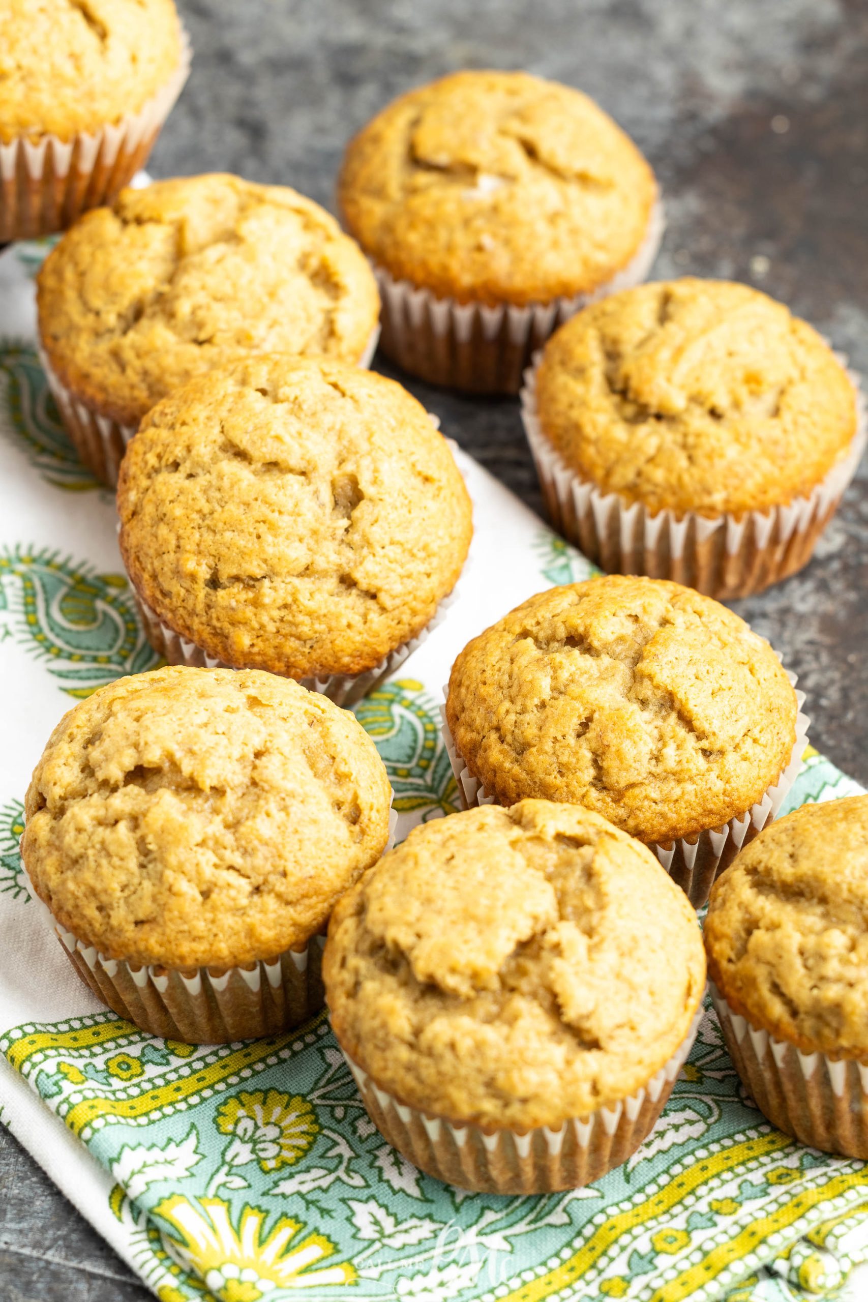 Eight plain muffins in paper liners are arranged on a patterned green and white cloth, set on a dark, textured surface.