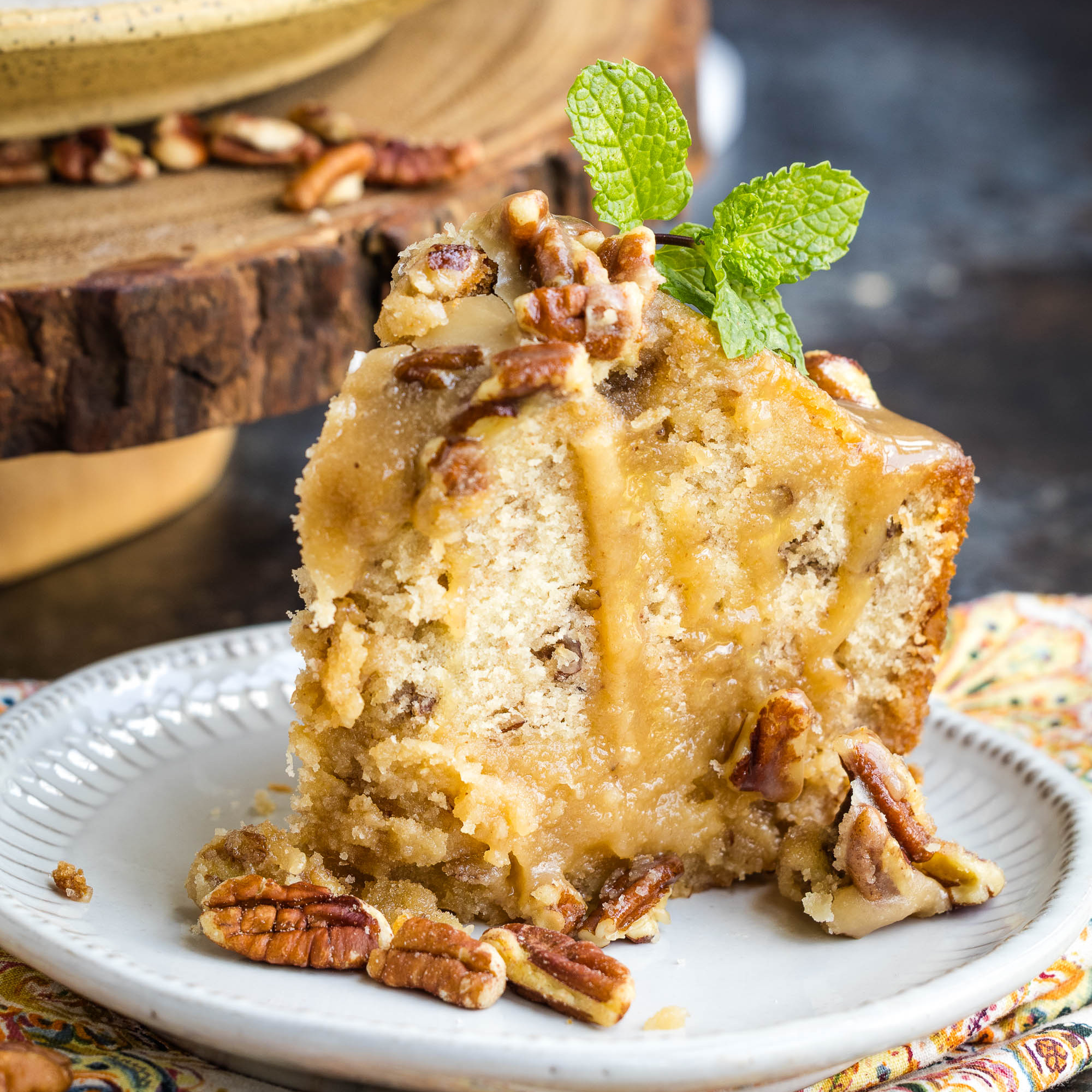 A slice of pecan cake with caramel glaze, topped with pecans and a sprig of mint, served on a white plate.