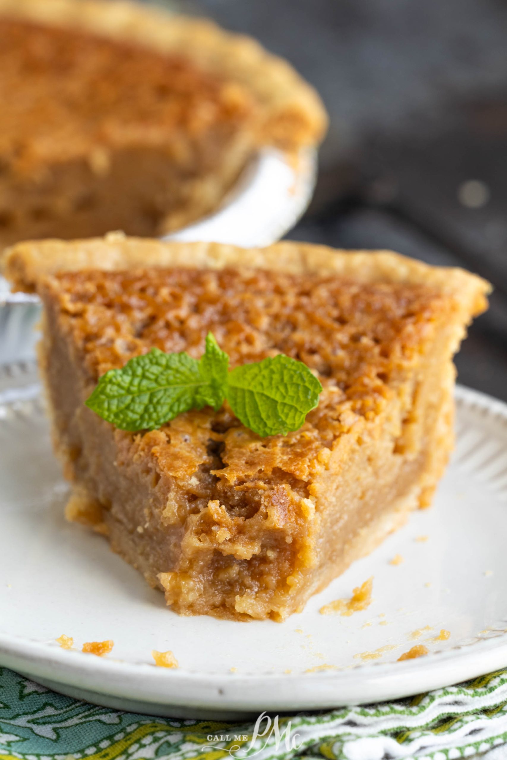 A slice of cookie butter chess pie garnished with a sprig of mint sits on a white plate, with the rest of the pie visible in the background.