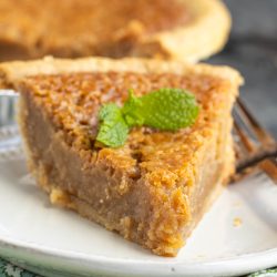 A slice of brown custard pie topped with a mint leaf sits on a white plate, with the rest of the pie visible in the background.