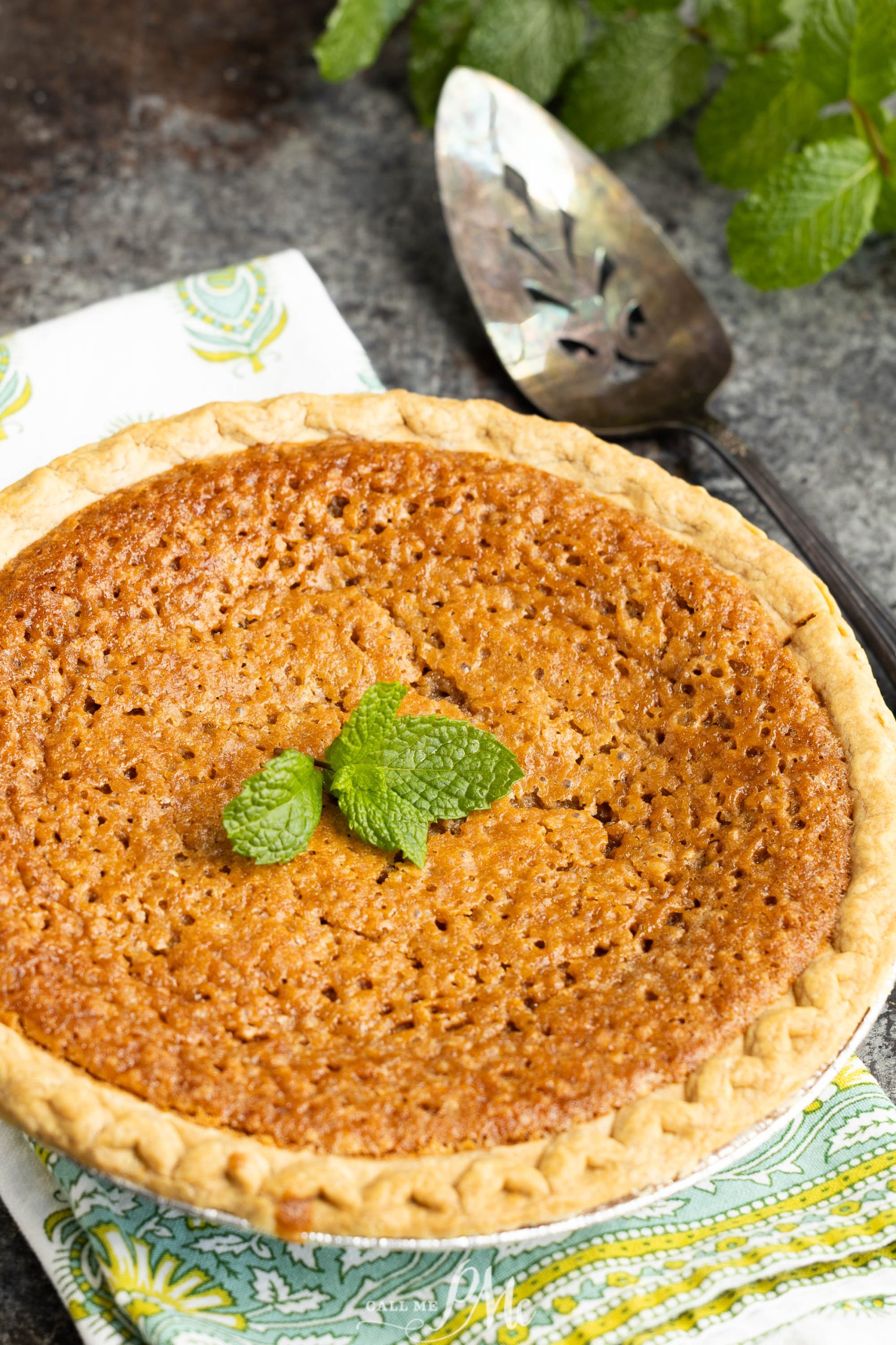 A cookie butter chess pie with a golden-brown filling, garnished with mint leaves, sits on a patterned cloth next to a metal pie server.