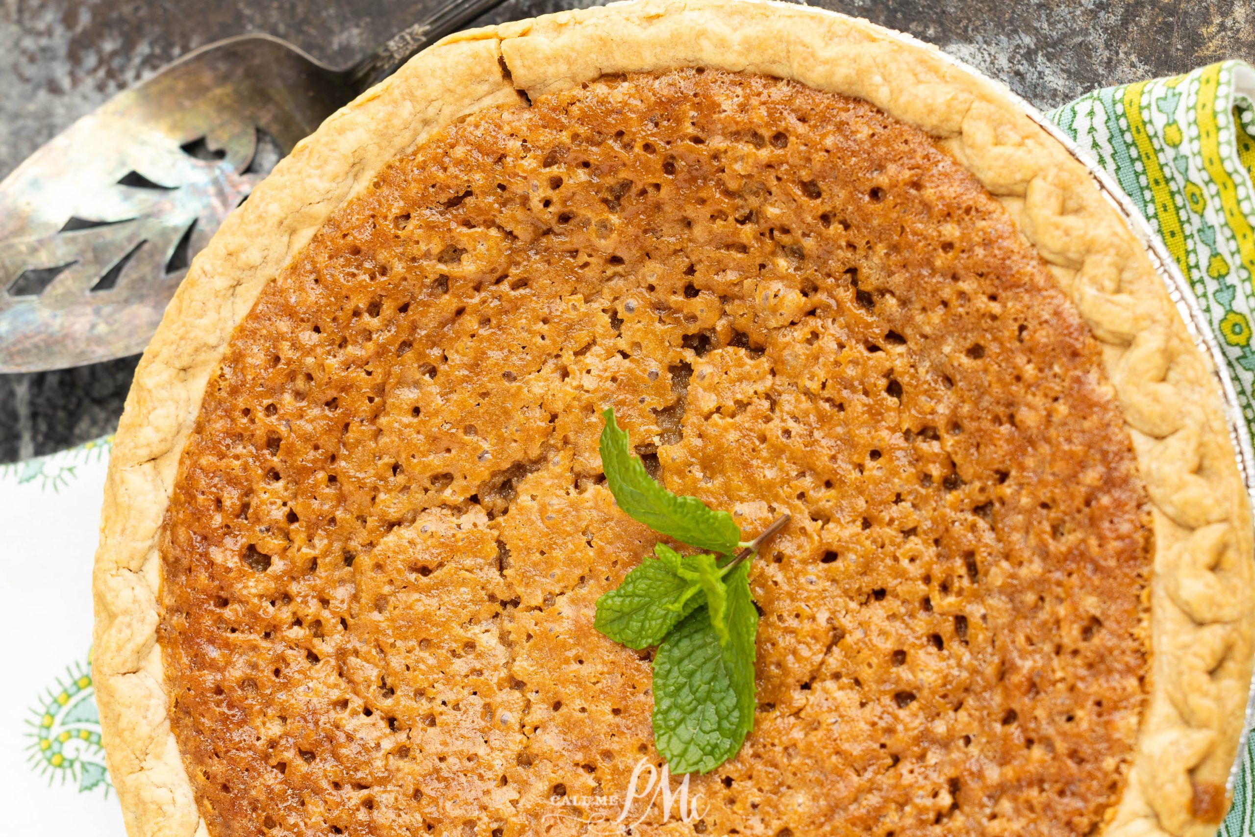 Close-up of a baked pie with a golden, textured crust and filling, garnished with a sprig of mint, placed next to a metal server on a patterned cloth.