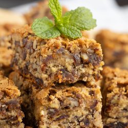 Close-up of stacked pecan praline bars topped with a sprig of fresh mint, surrounded by scattered pecan halves on a wooden surface.