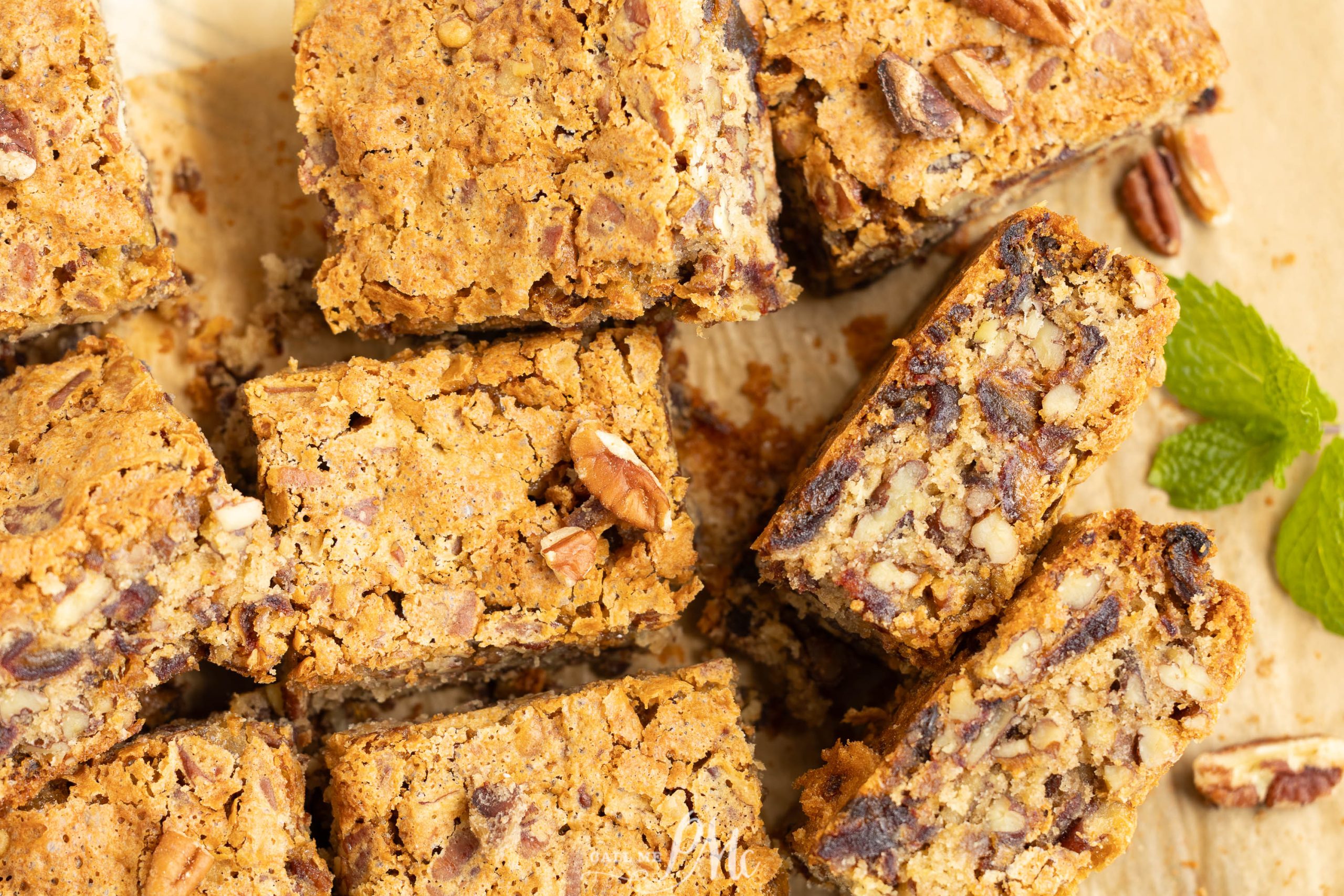 Close-up of several pecan date bars, some stacked and some lying flat, on parchment paper with a sprig of mint nearby.