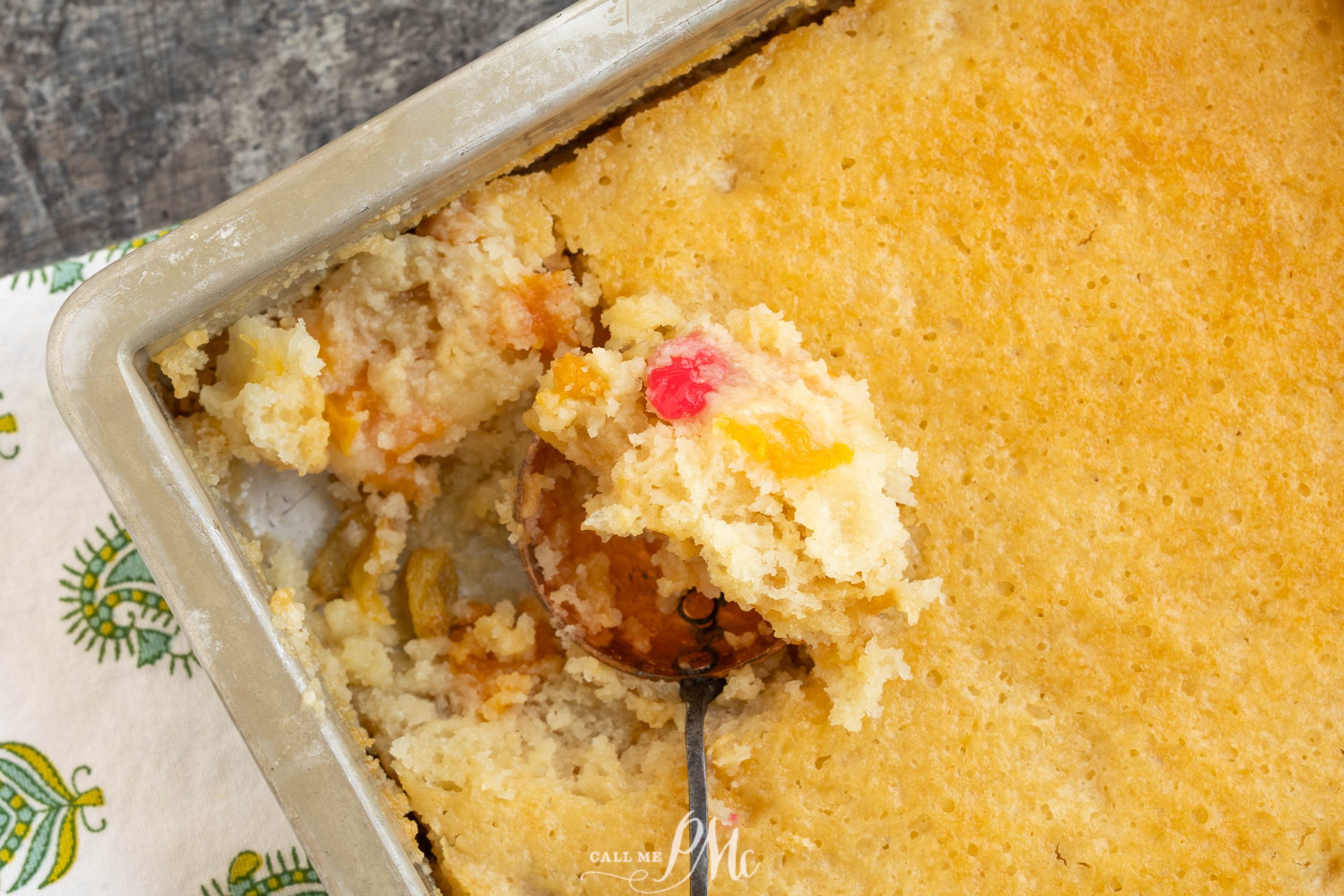 A close-up of a baking pan with a serving of peach cobbler being scooped out, showing a golden crust and visible pieces of peach and a cherry.