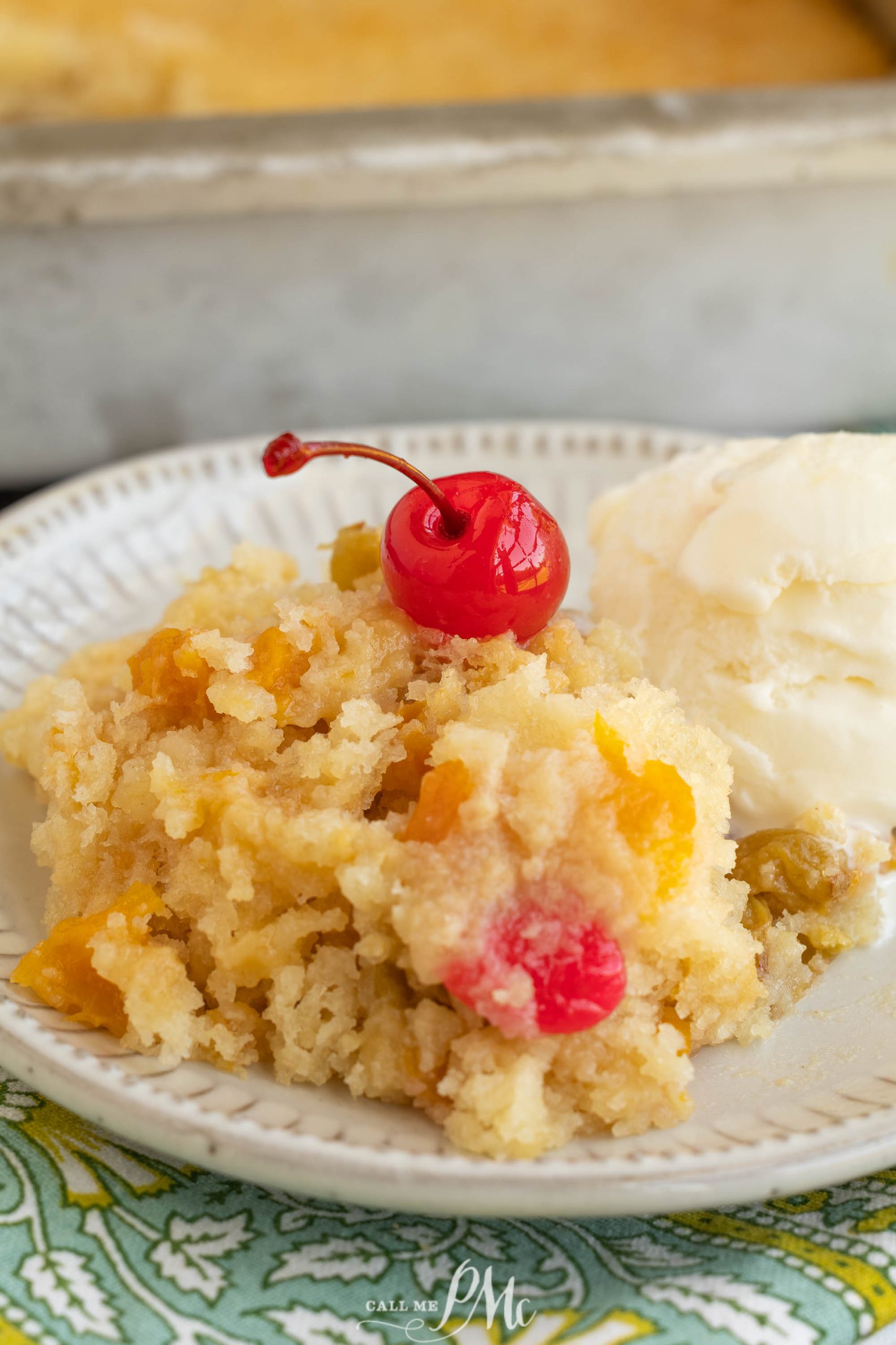 A serving of cobbler with visible fruit pieces and a maraschino cherry on top, alongside a scoop of vanilla ice cream on a white plate.
