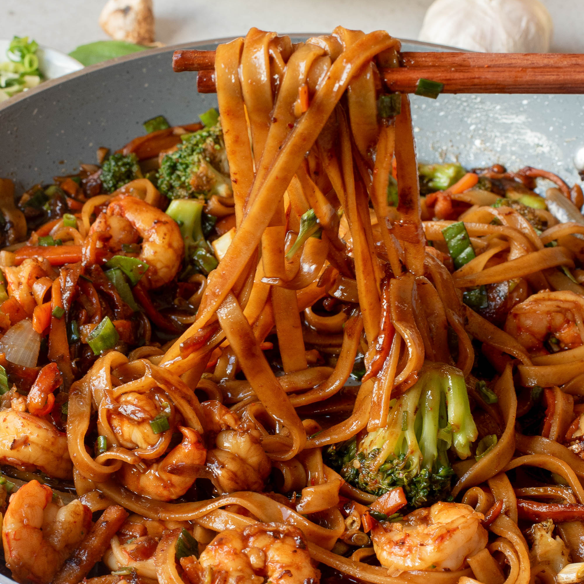 A close-up of stir-fried noodles with shrimp, broccoli, and vegetables in a pan, with chopsticks lifting a portion.