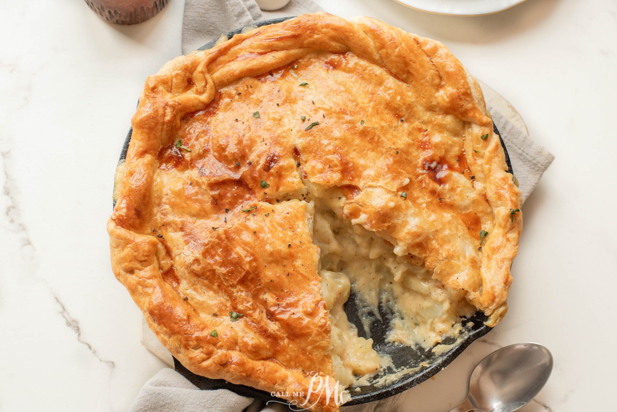 A golden-brown pot pie in a round baking dish with a slice removed, revealing a creamy filling. A spoon lies nearby on a white surface.
