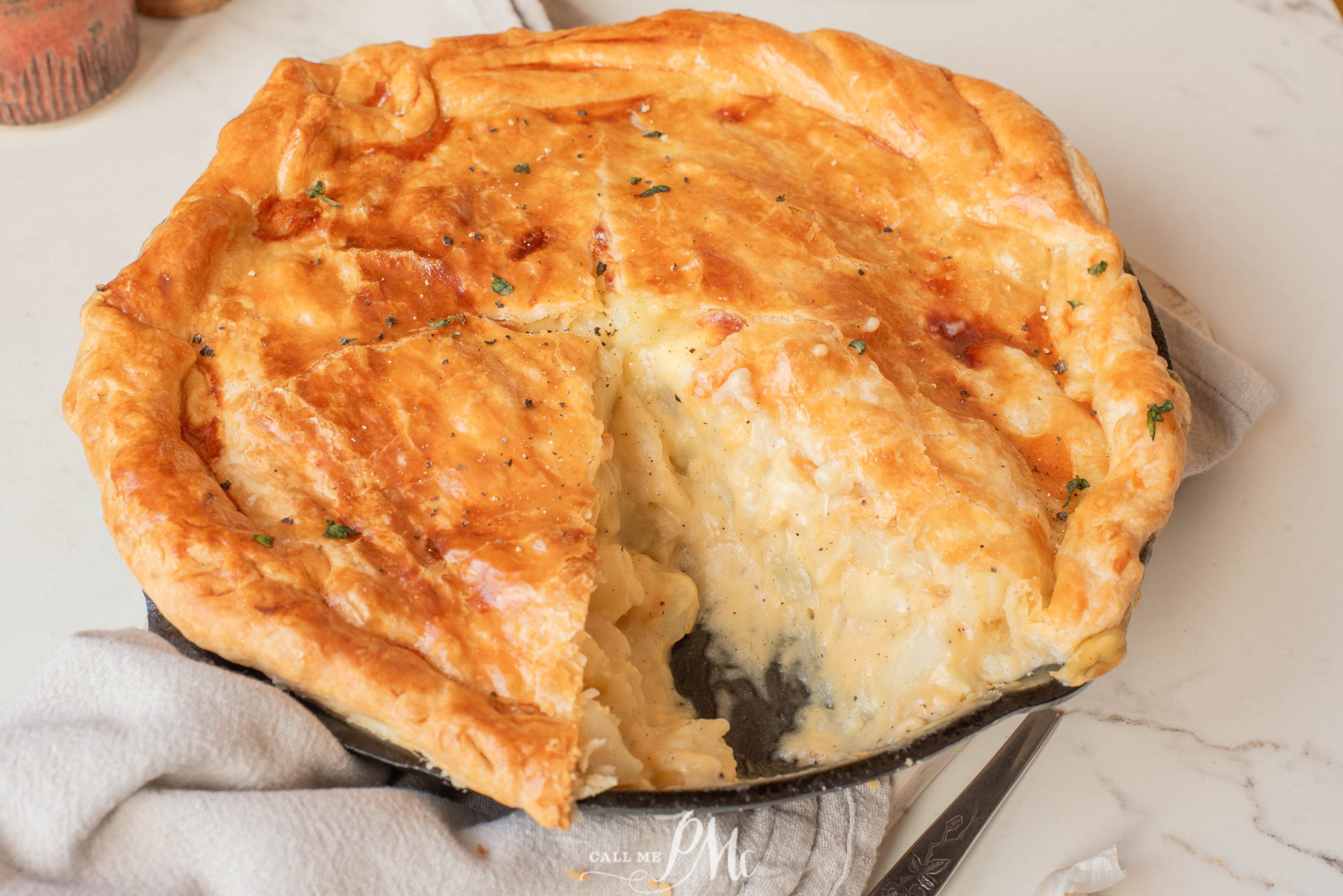 A golden-brown pot pie with a large slice removed, showing a creamy filling inside, sits on a white surface.