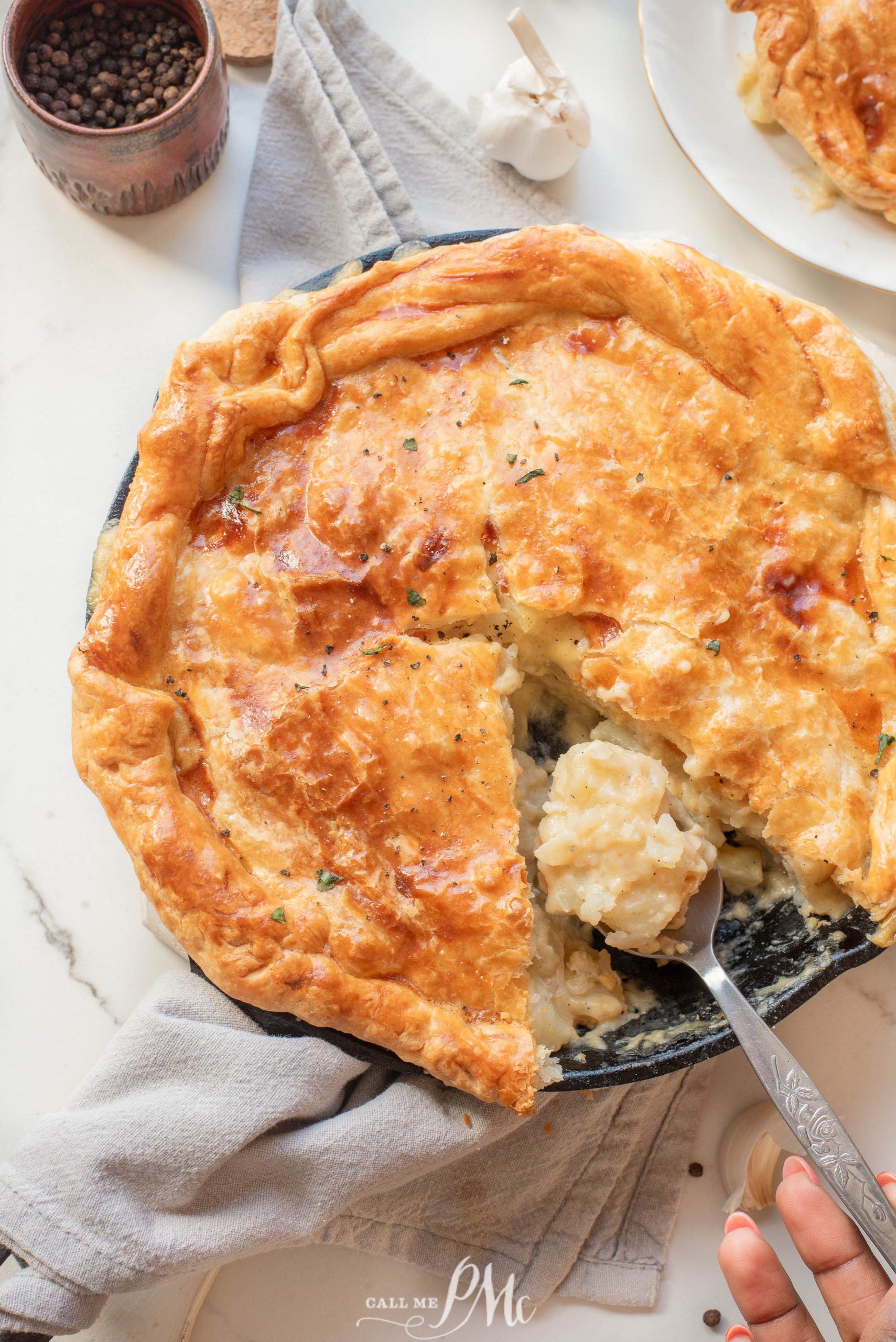 Potato Cheese Onion Pot Pie with a flaky crust in a black dish, a slice being served with a spoon, set on a light surface with a napkin and seasoning around.