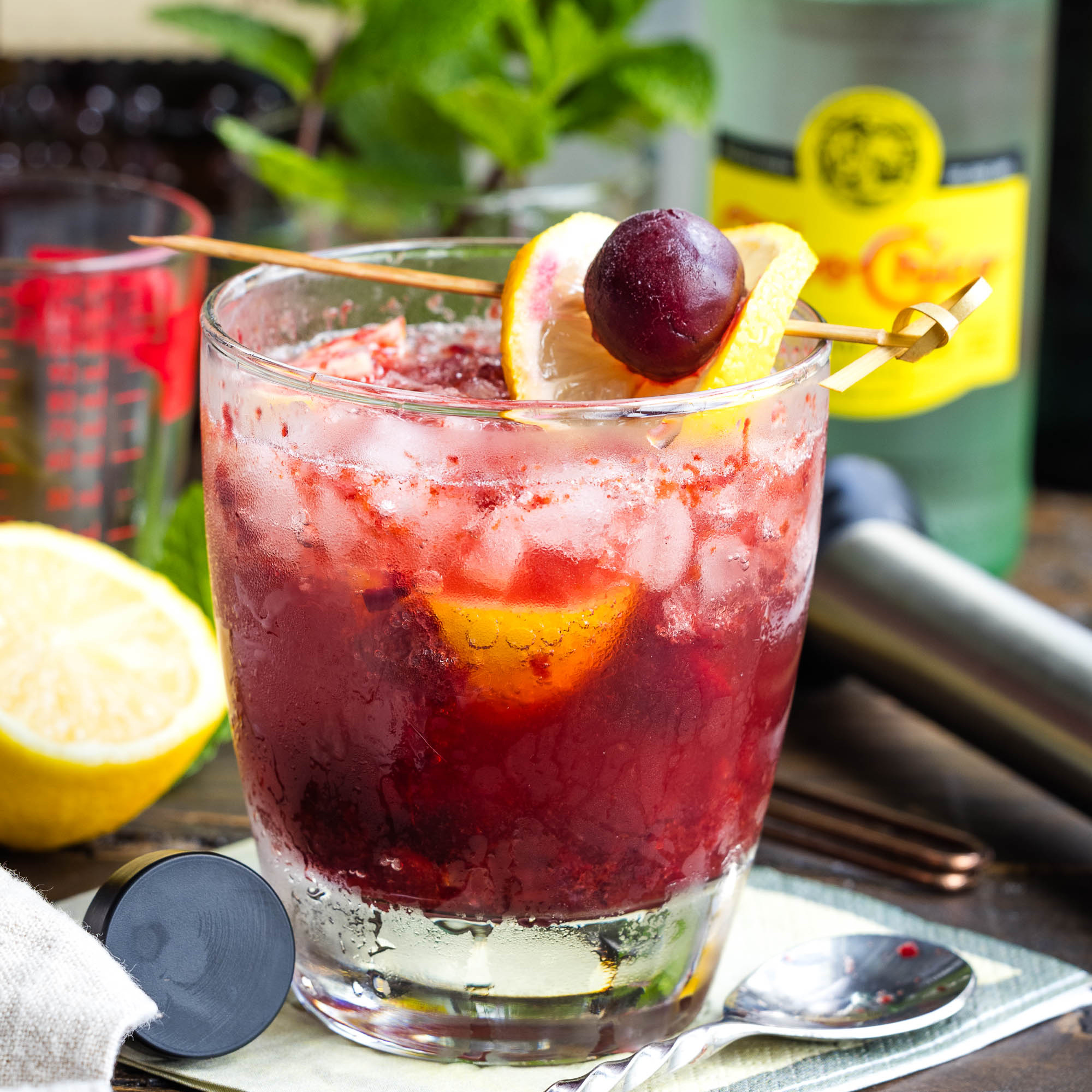 A glass of iced cherry cocktail garnished with a lemon slice and cherry, with a lemon half, mint, and a bottle in the background.