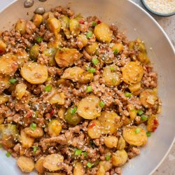 A stainless steel pan filled with stir-fried Brussels sprouts, ground meat, and diced vegetables, garnished with sesame seeds and chopped green onions.