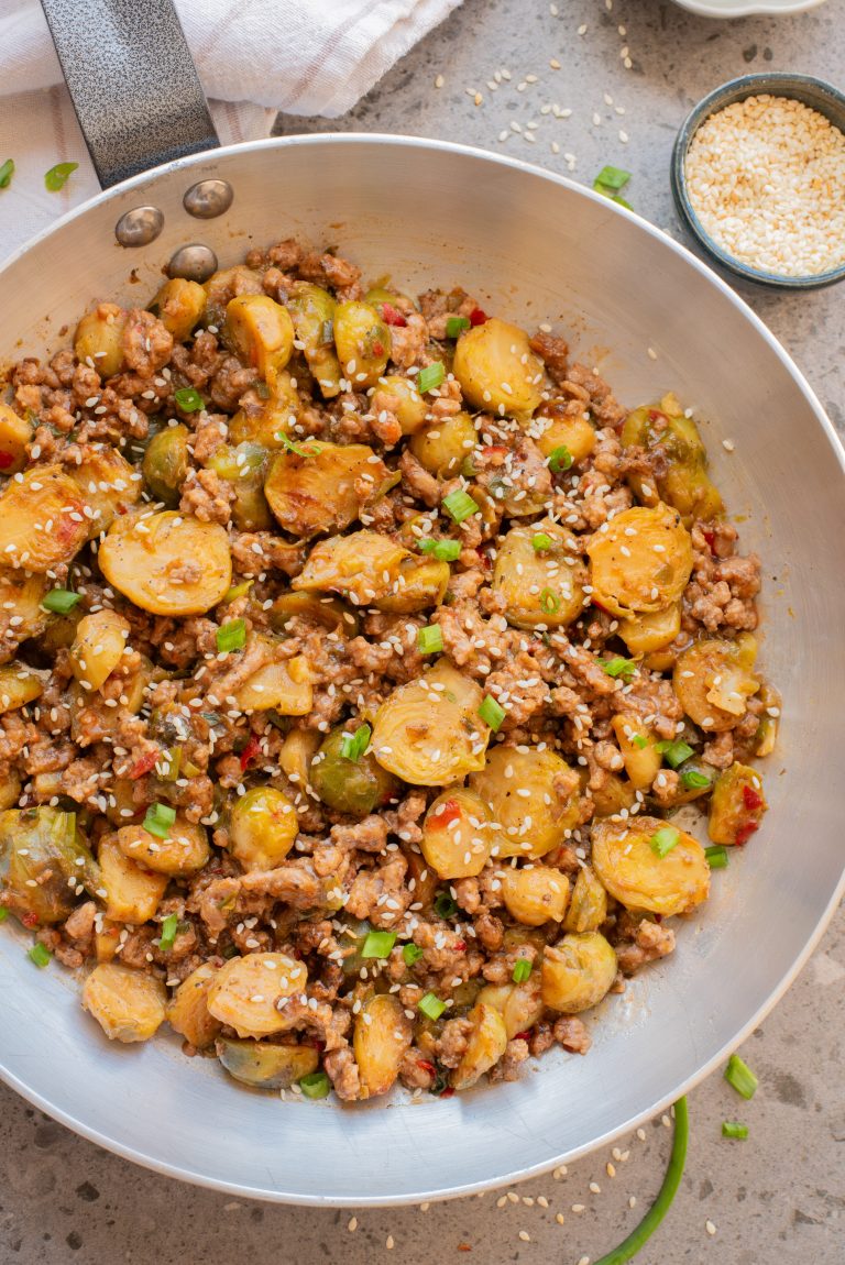 A stainless steel pan filled with stir-fried Brussels sprouts, ground meat, and diced vegetables, garnished with sesame seeds and chopped green onions.